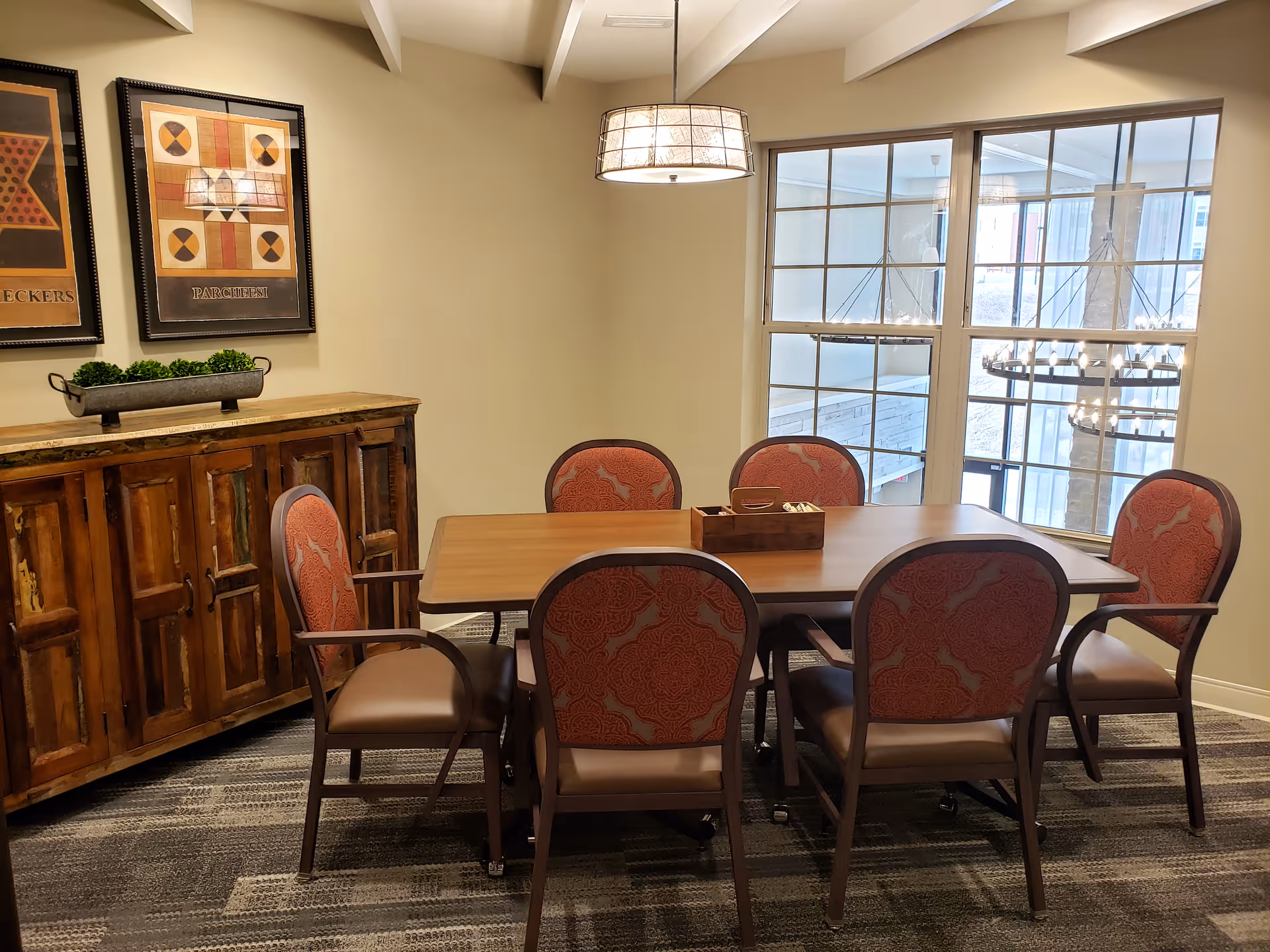 A dining room with a wooden table surrounded by six upholstered chairs with red patterned backs and brown seats. A wooden sideboard with a decorative plant is against the wall, which has two framed game-themed artworks. A large window lets in natural light, and a modern hanging light fixture is above the table.