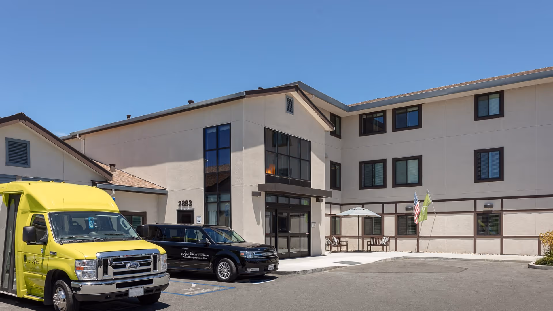 Exterior view of a three-story senior living facility building with beige walls and brown trim under a clear blue sky. There is a yellow shuttle bus and a black vehicle parked in front near the entrance, which has large glass doors and windows. Two flags are displayed near a small seating area with an umbrella.
