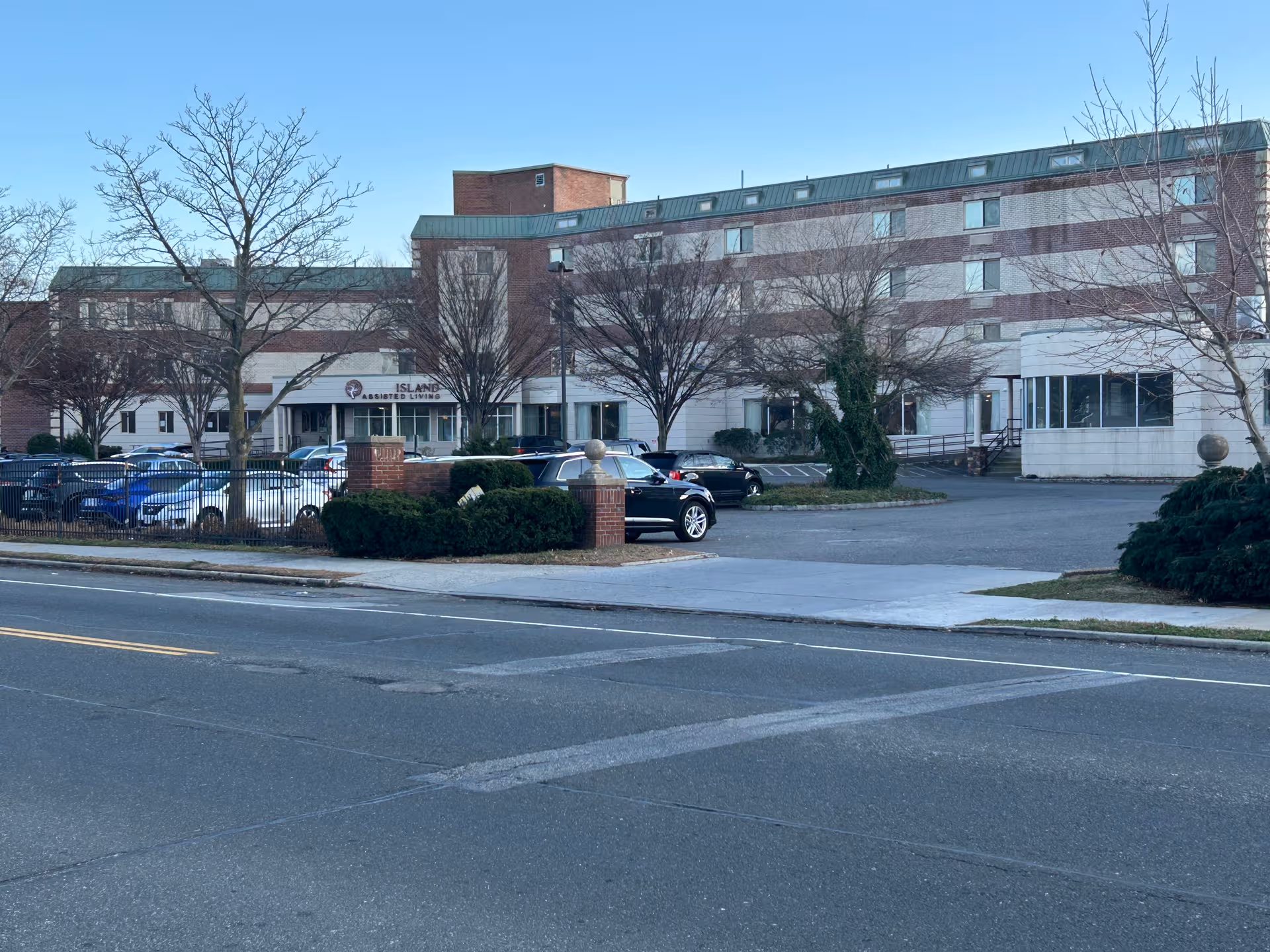 Brick nursing home building with cars parked in front and leafless trees along the street.