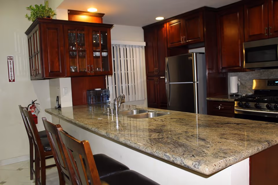Modern kitchen with a large granite island and sink, bar stools, dark wood cabinets, and stainless steel refrigerator and stove.