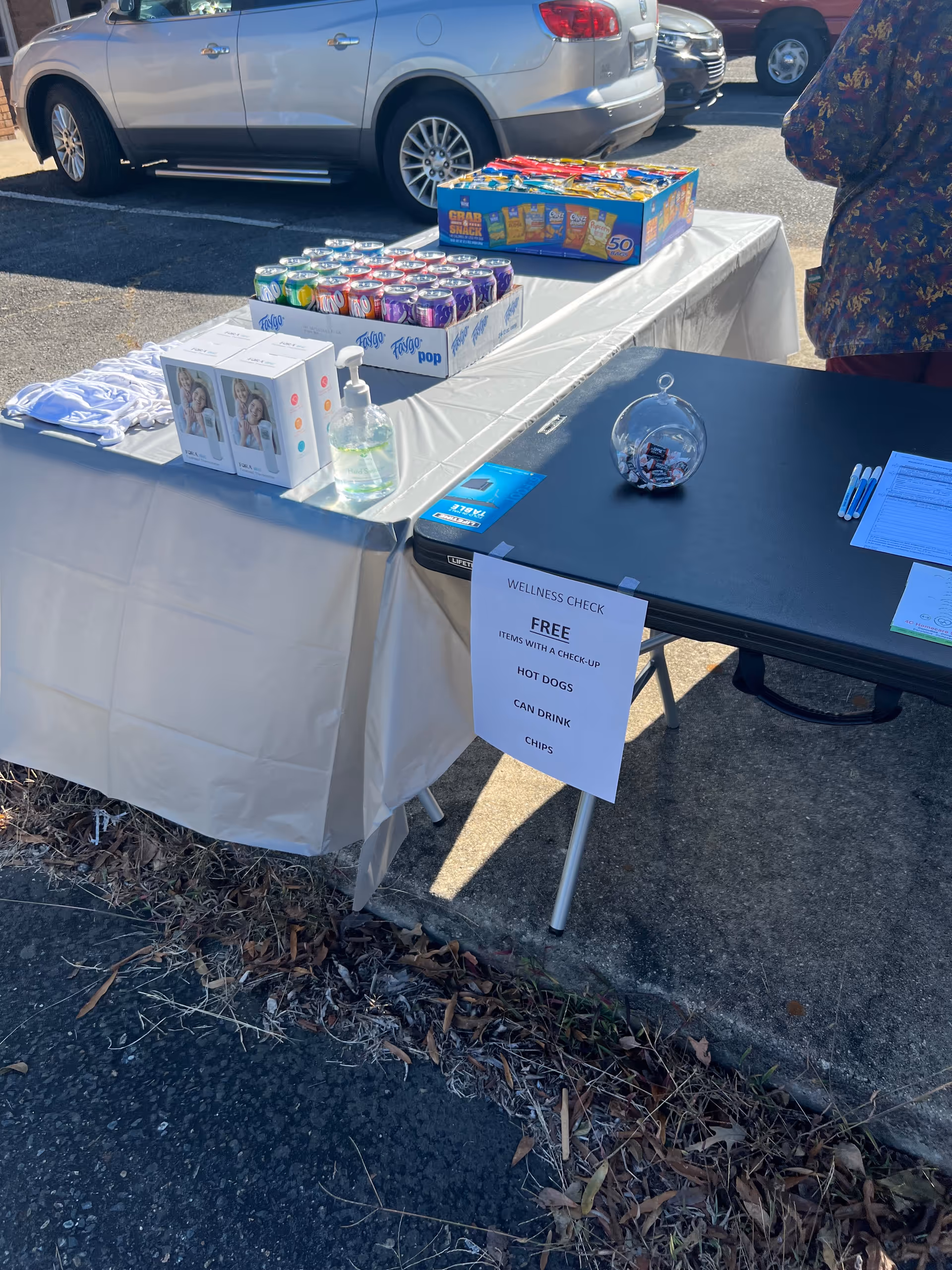 Outdoor table with snacks, drinks, hand sanitizer, and a wellness sign at a parking lot event.