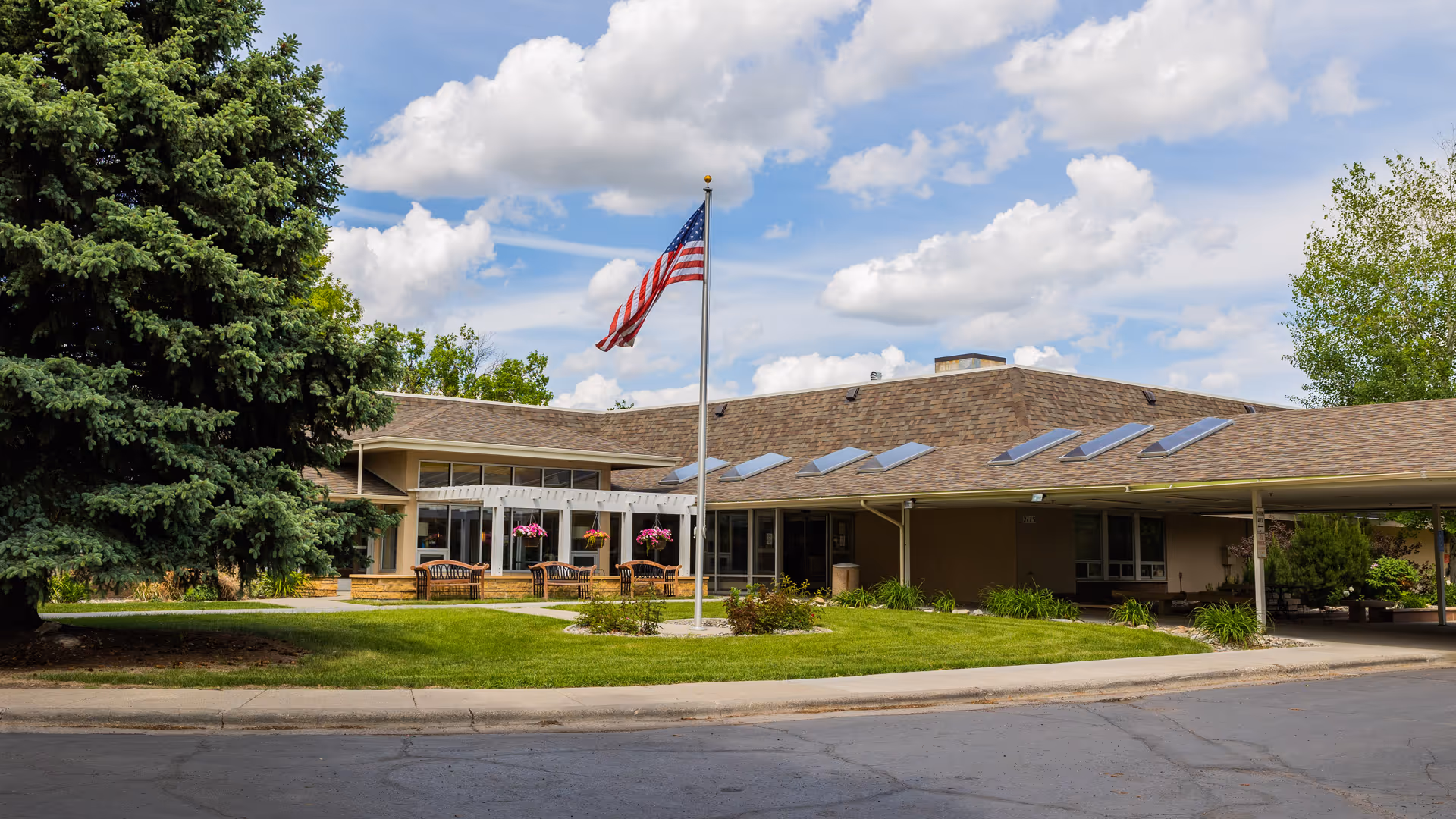 Front entrance of a low-rise nursing facility with an American flag, benches, hanging flower baskets, and a manicured lawn under a partly cloudy sky.