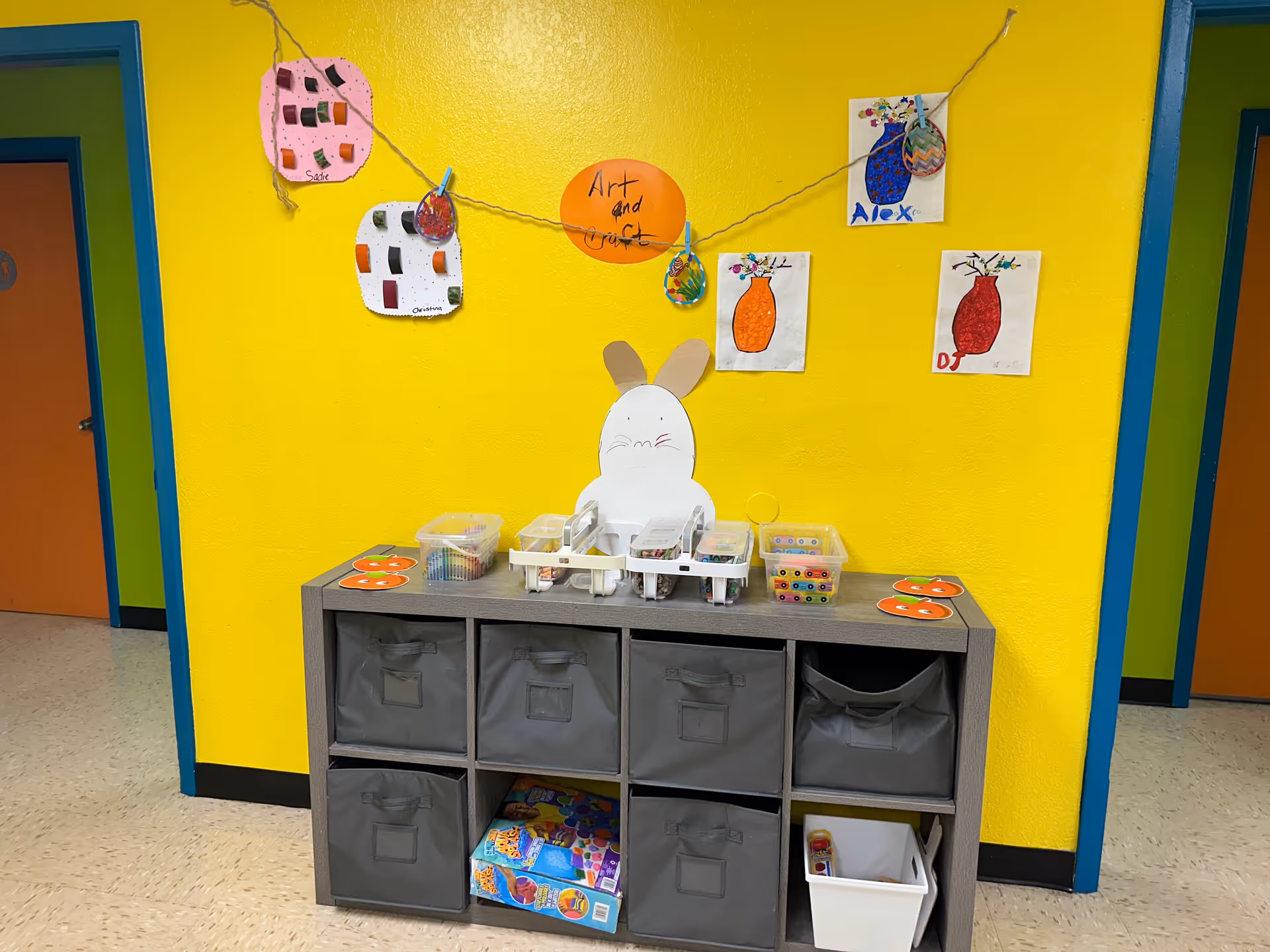 Bright yellow wall with children's art, bunny cutout, and cubby storage in a colorful interior.