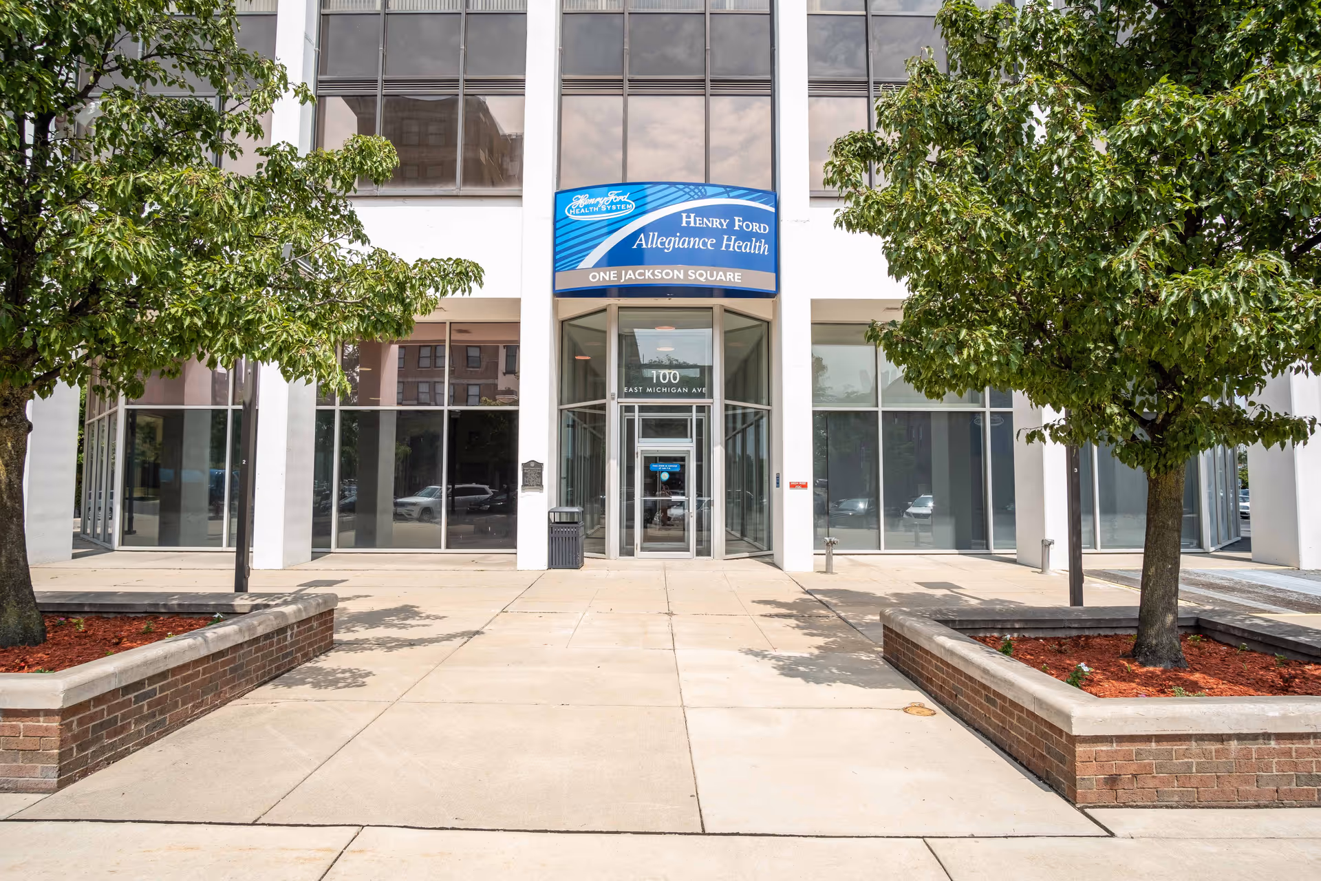Front entrance of a modern glass-front building with trees and planters.