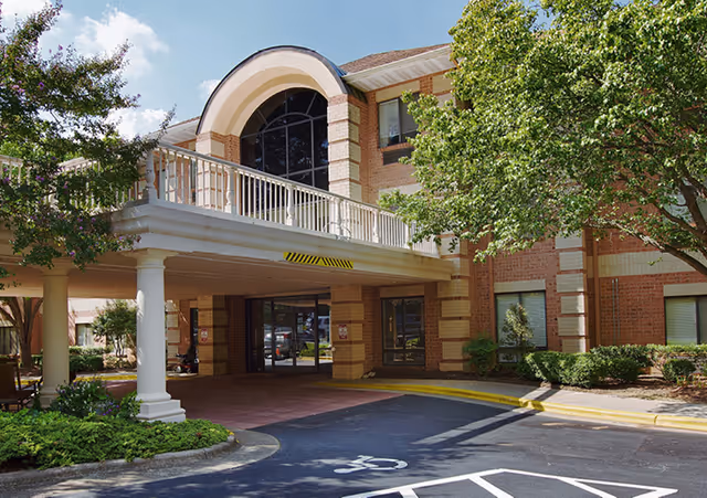 Exterior view of a brick building with a covered entrance supported by white columns. There are trees and shrubs around the entrance area, and a driveway with a handicapped parking space is visible in front.