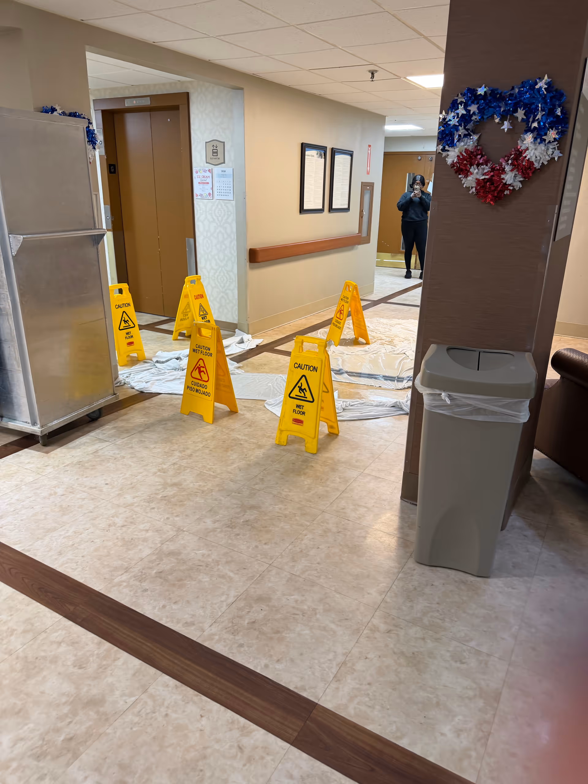 Indoor hallway with wet floor signs, a trash can, and a person taking a photo.