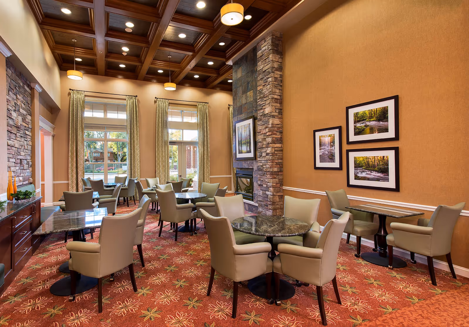 A spacious and elegant dining room with multiple round and rectangular tables surrounded by beige upholstered chairs. The room features a high coffered ceiling with recessed lighting and hanging pendant lights. Large windows with patterned curtains allow natural light to fill the space. A stone fireplace is built into one wall, which is decorated with framed nature photographs. The carpet has a floral pattern in warm tones.