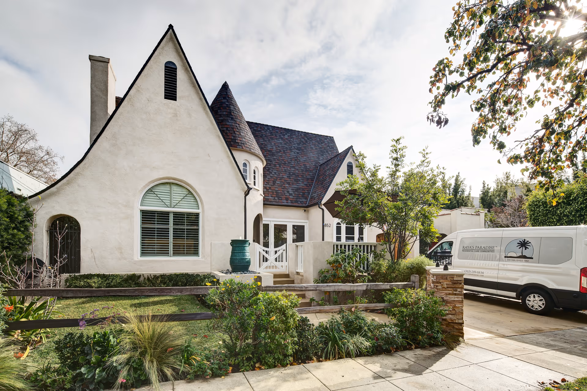 White stucco senior care building with front yard, gate, and Raya's Paradise van.