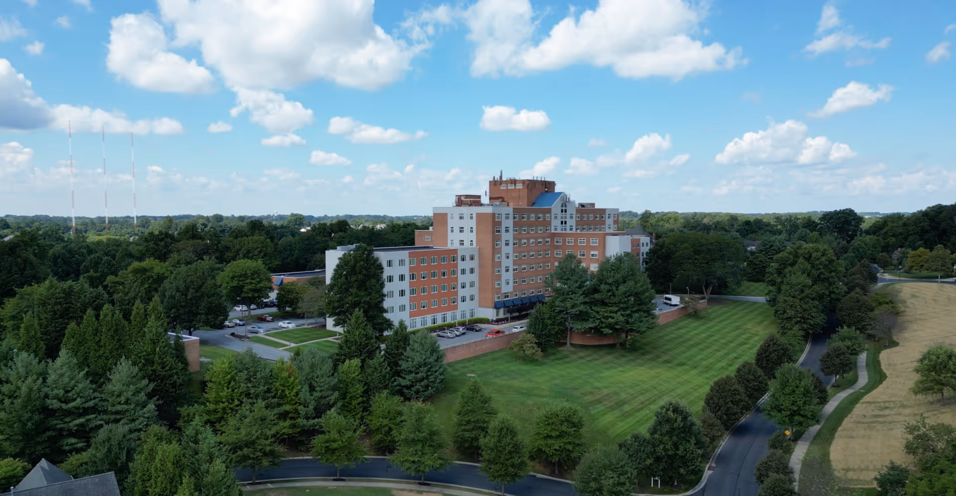Aerial view of a multi-story senior living building amid trees, parking, and expansive green grounds.