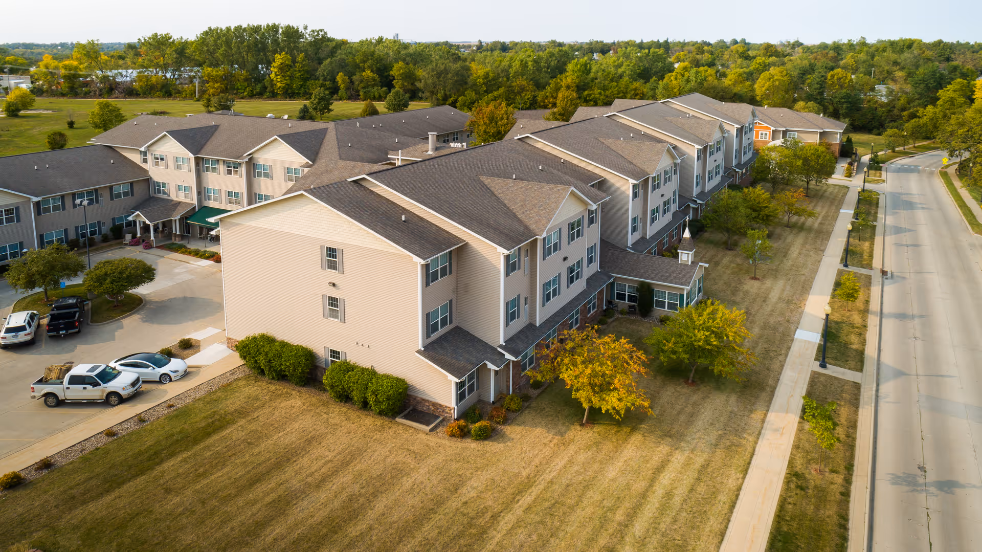 Aerial view of a large, multi-story senior living facility building with beige siding and a gray roof, surrounded by well-maintained lawns, trees, and a parking area with several vehicles. A road runs alongside the property, and there is a backdrop of dense green trees in the distance.