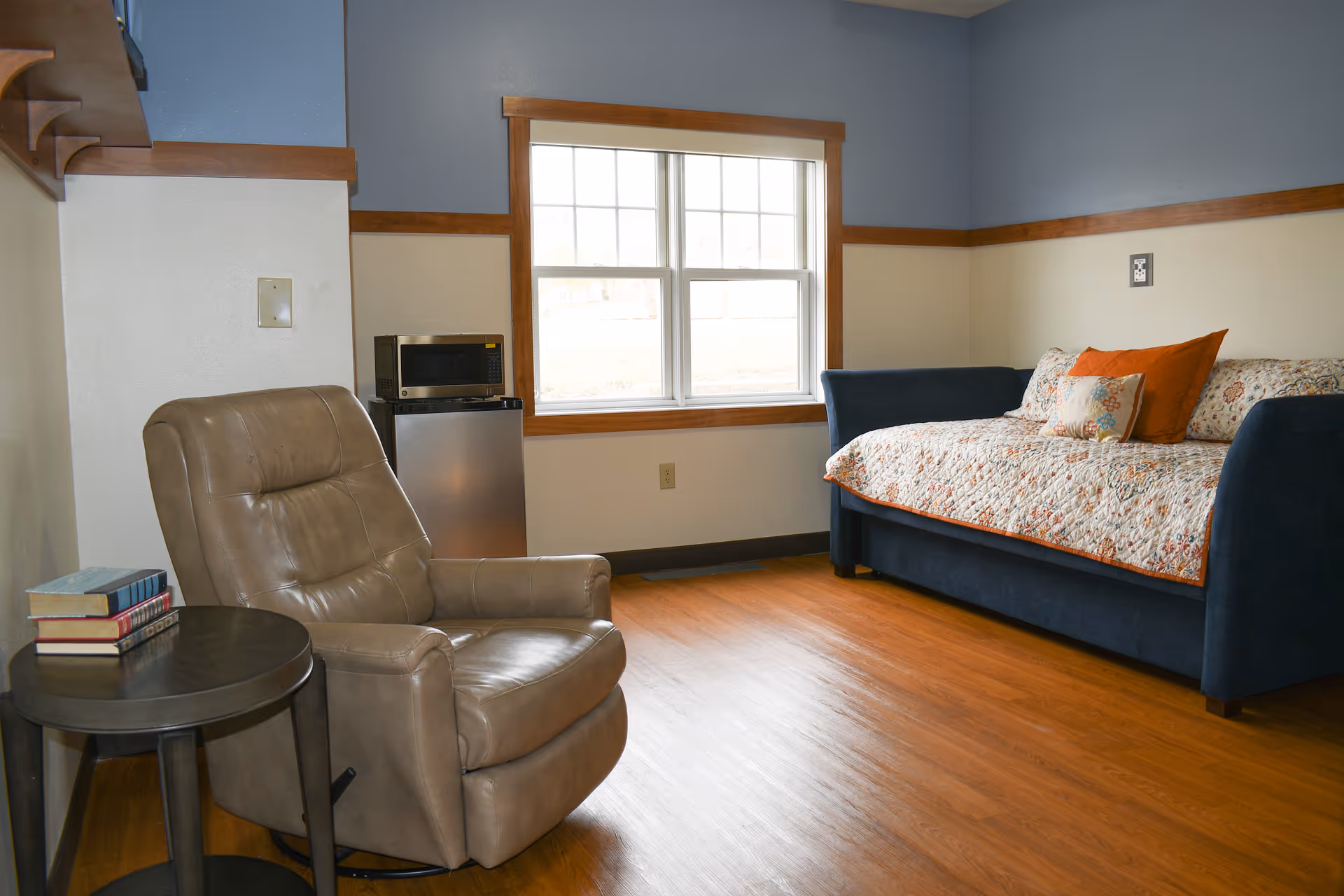 A cozy respite suite living area with a chair, sofa bed, window, and wood floors.
