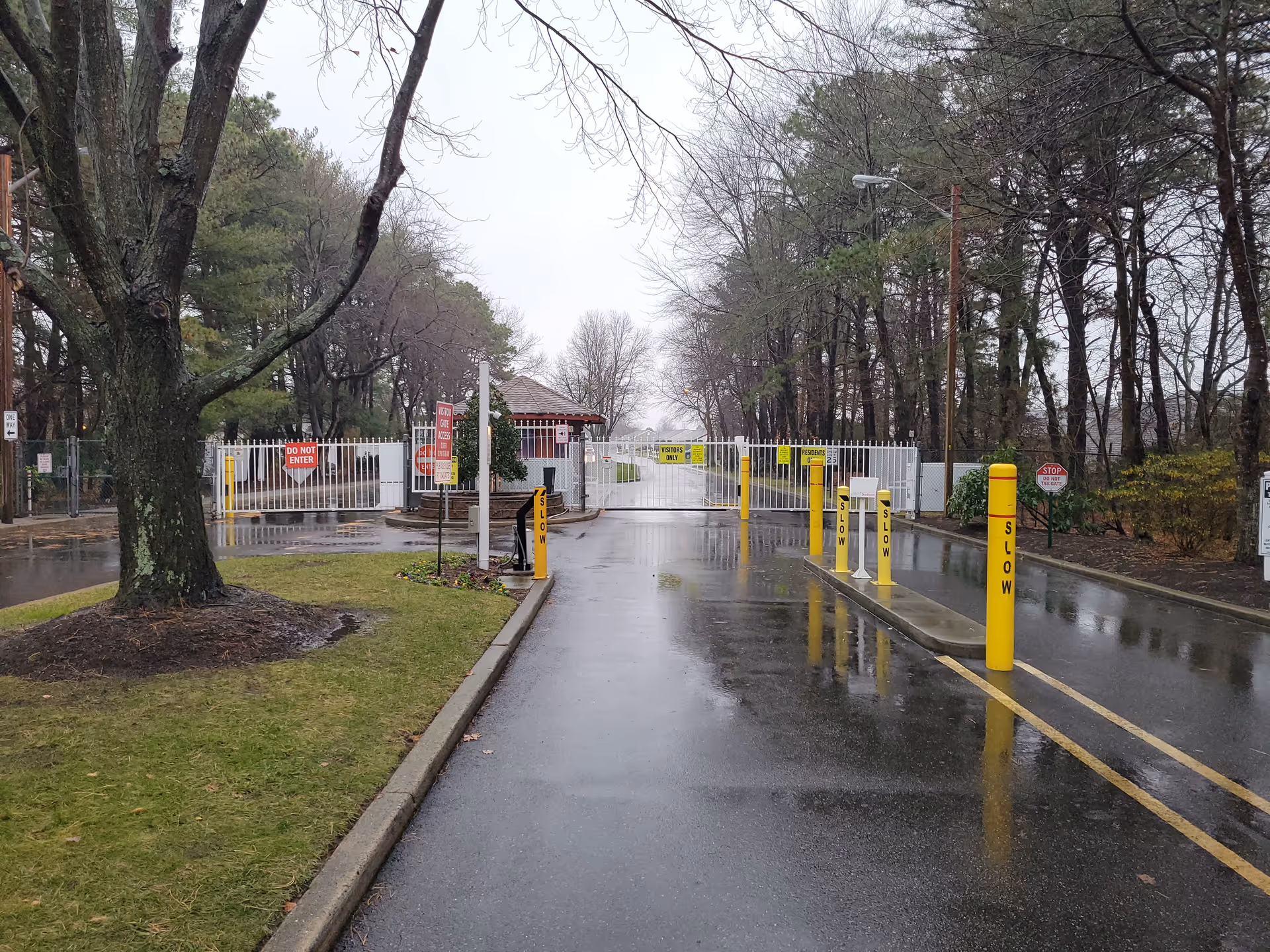 Gated exterior entrance to Leisure Knoll Manchester with rainy road, yellow posts, and trees.
