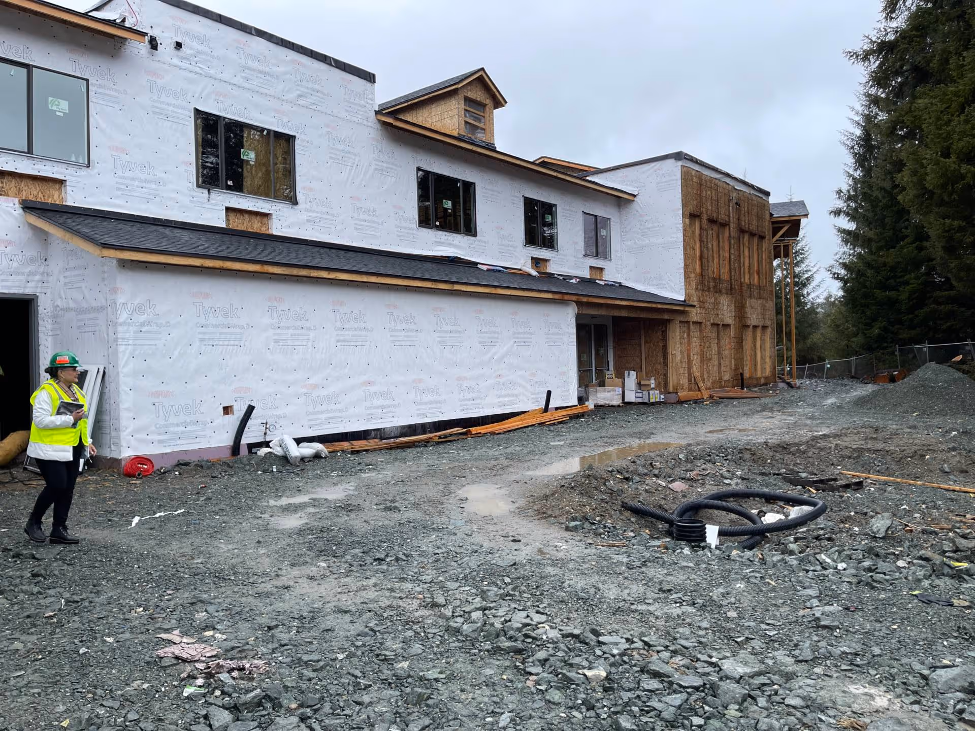 Front exterior of a multi-story senior living building under construction with Tyvek wrap, construction materials on a gravel lot, and a worker in a high-visibility vest.