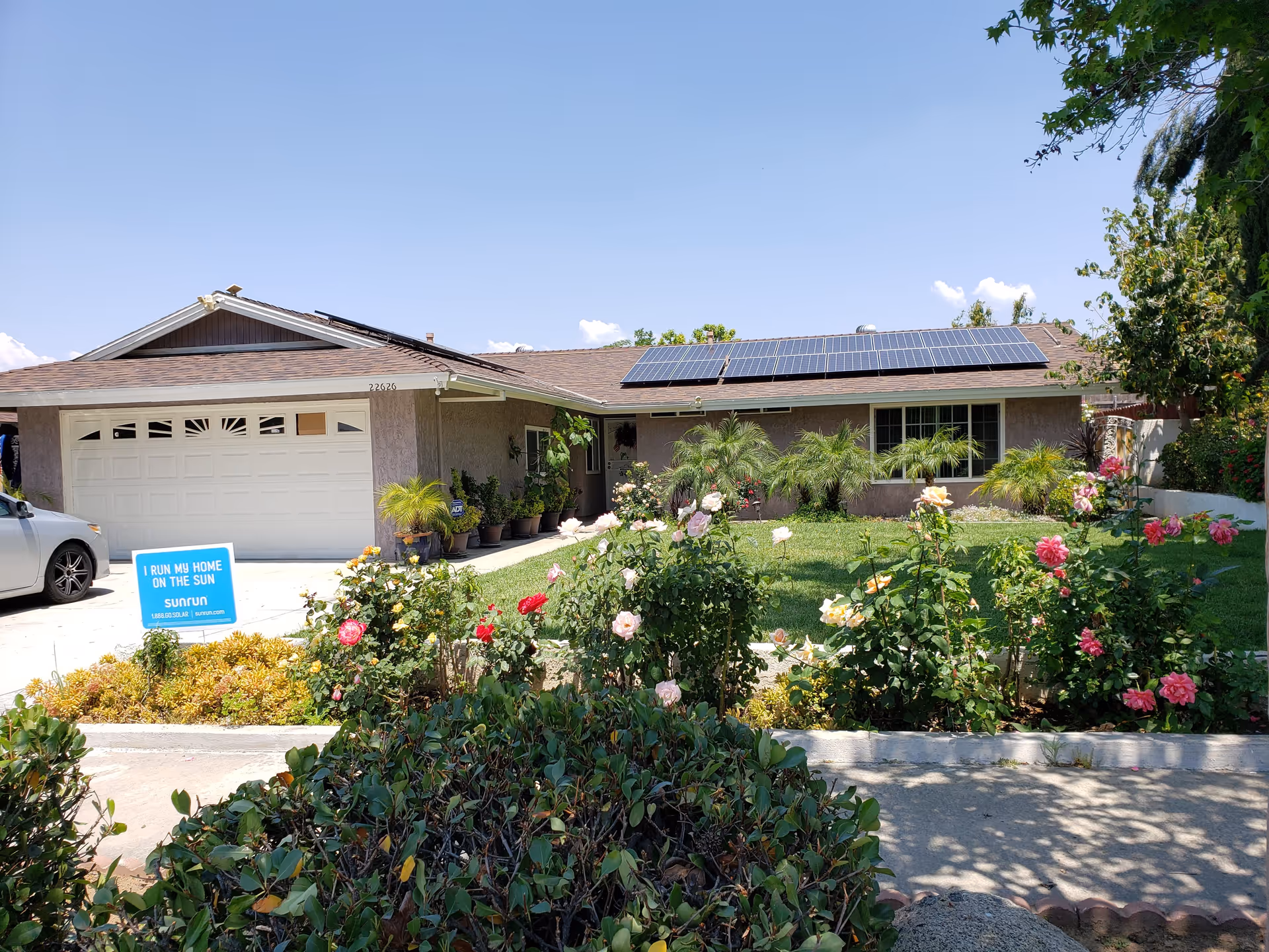 Single-story house with a brown roof equipped with solar panels, a white garage door, and a driveway with a parked silver car. The front yard features a well-maintained green lawn, various potted plants near the entrance, and a garden bed with blooming roses in multiple colors. A blue sign in the yard reads 'I RUN MY HOME ON THE SUN sunrun 1.888.60.SOLAR sunrun.com'.