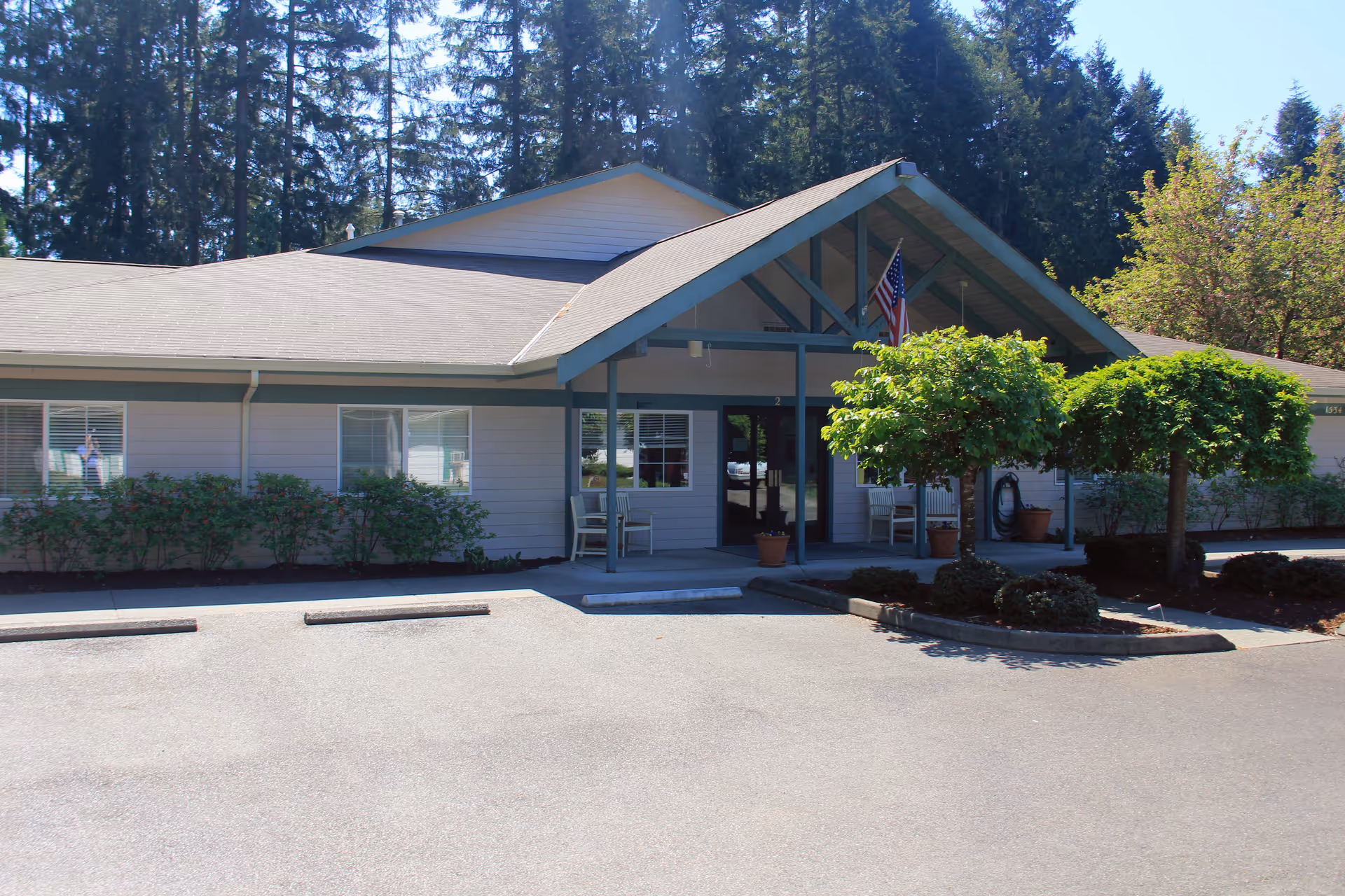 Exterior view of a single-story building with a covered entrance supported by beams. There are two small trees and some bushes in front of the building, along with a few chairs on the porch. An American flag is hanging near the entrance. The building is surrounded by tall trees and a paved parking area is visible in the foreground.