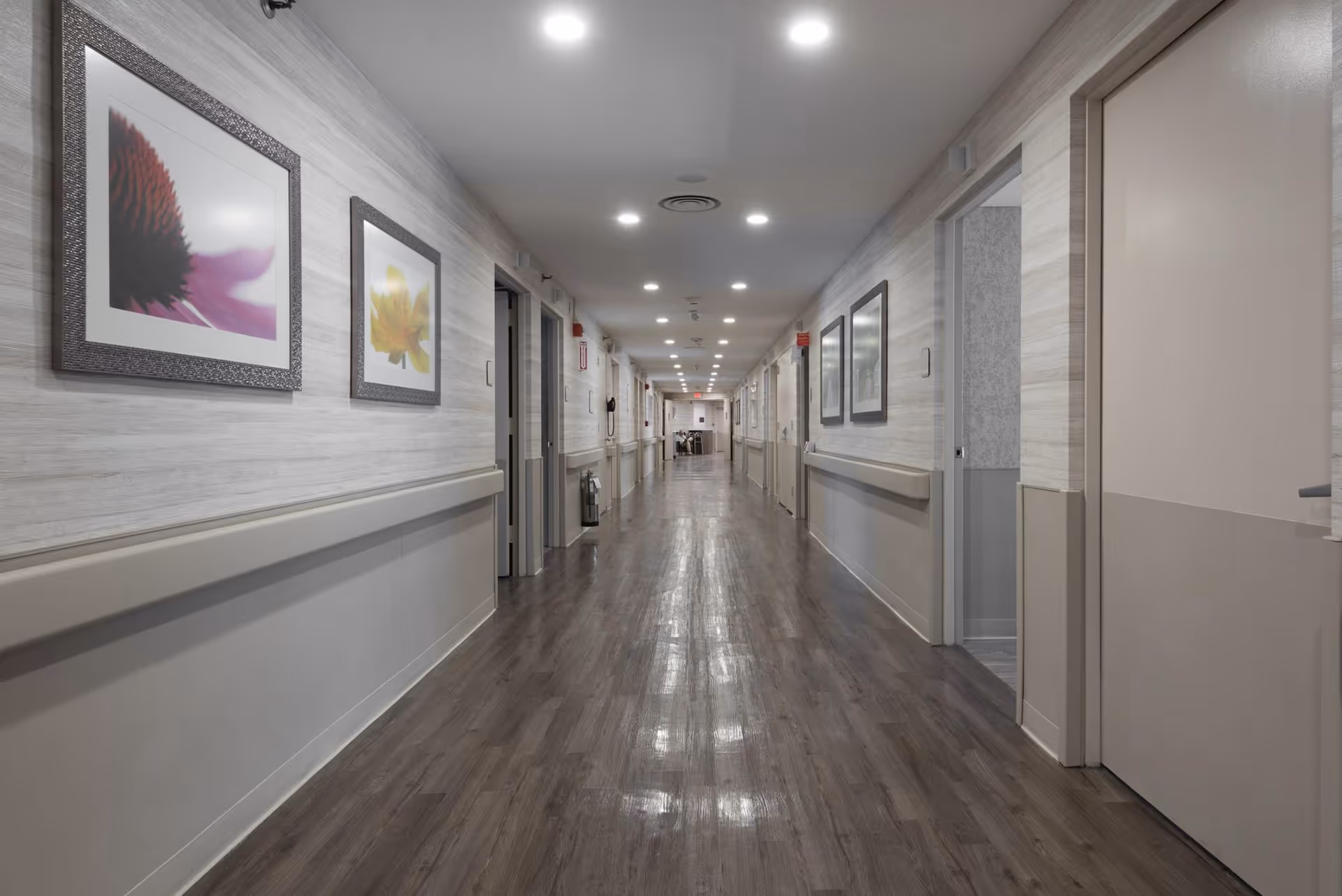 Long, well-lit interior hallway in a senior living facility with framed artwork on the walls and doors to rooms on both sides.