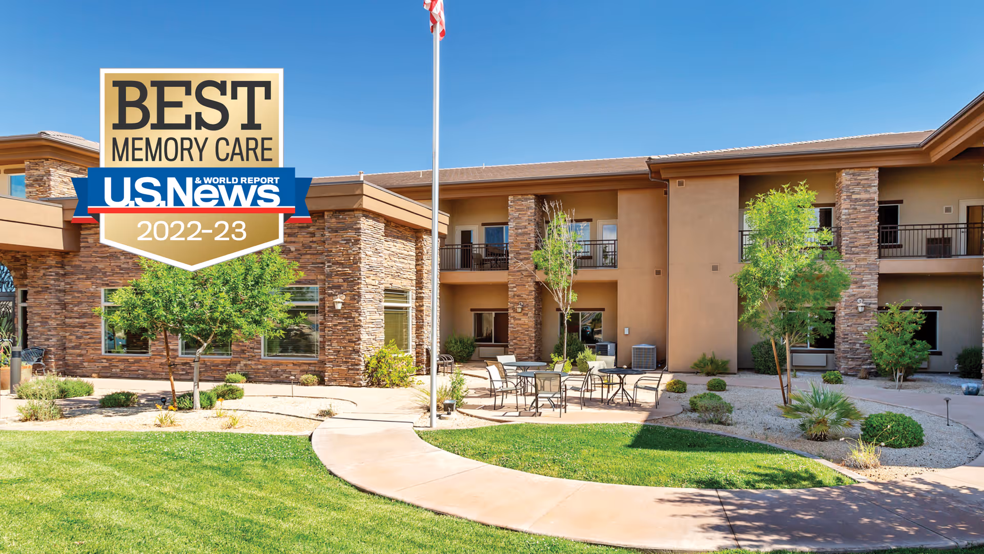 Courtyard and front exterior of a two-story senior living building with patio seating, desert landscaping, and a prominent 'Best Memory Care U.S. News 2022-23' badge.