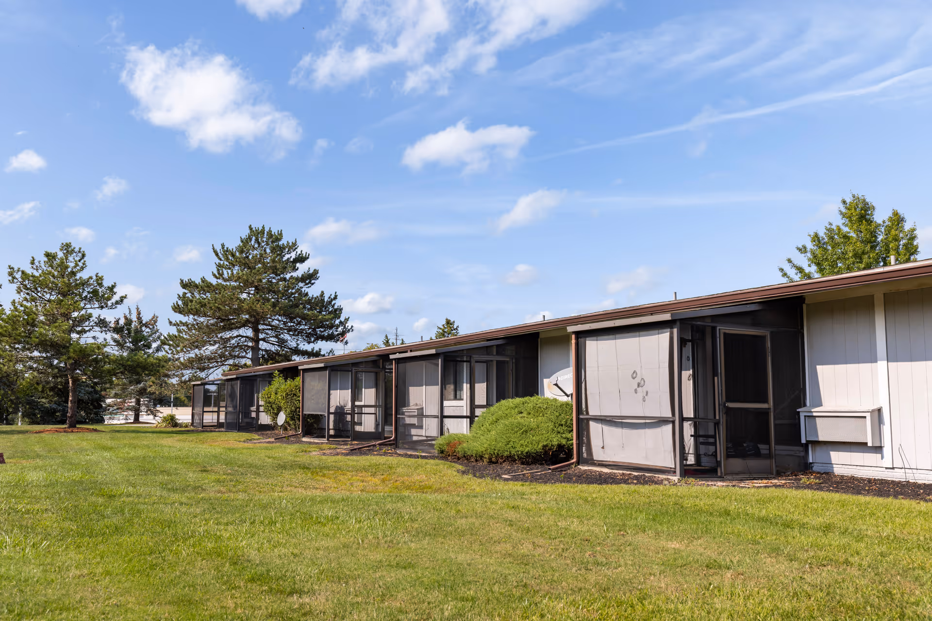 Single-story senior living building with screened-in patios facing a wide grassy lawn and trees under a blue sky.