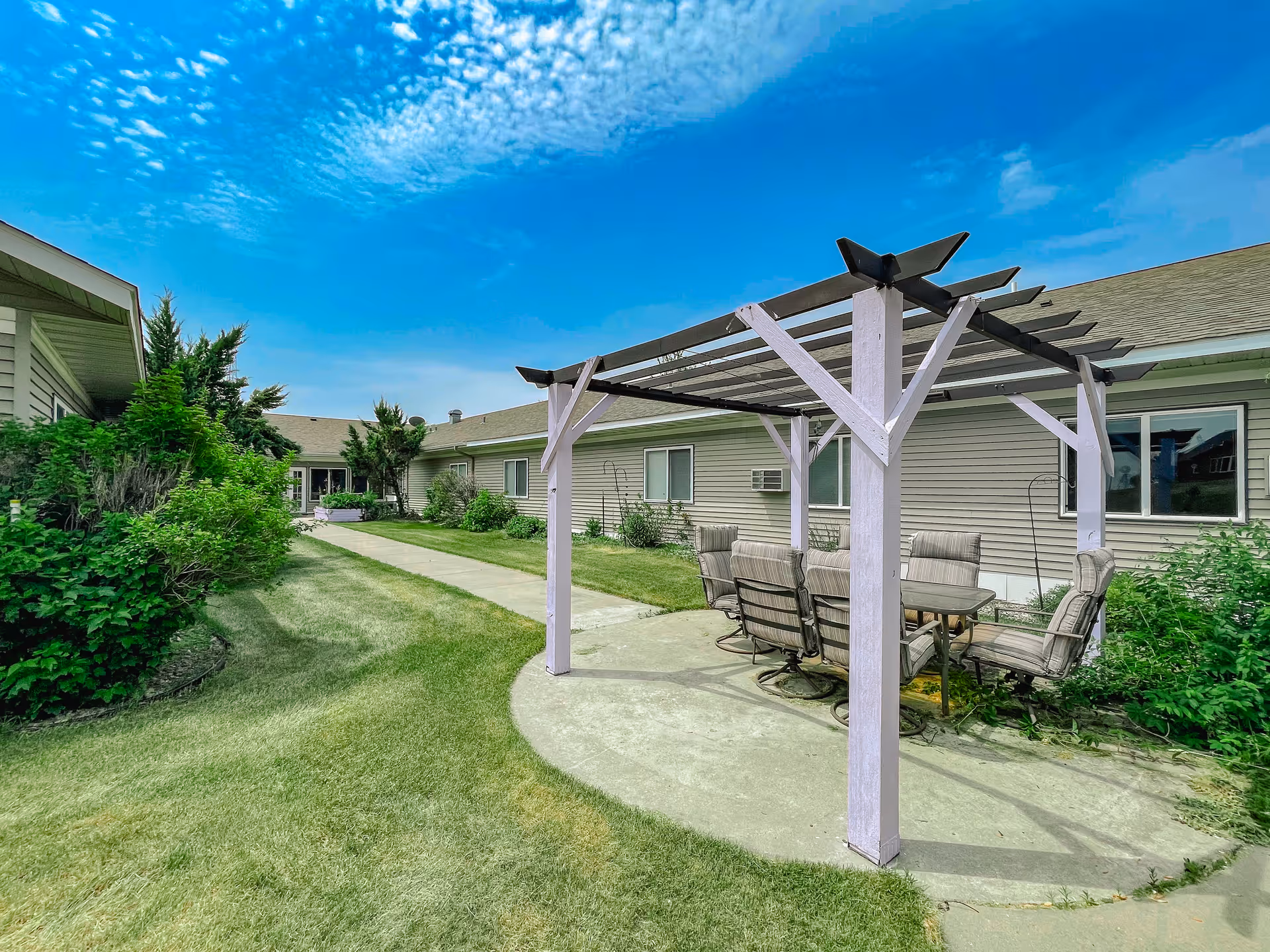 Outdoor patio area at Valley View Estates Assisted Living featuring a pergola with a glass-top table and cushioned chairs on a concrete slab, surrounded by green grass, bushes, and a building with beige siding under a partly cloudy blue sky.