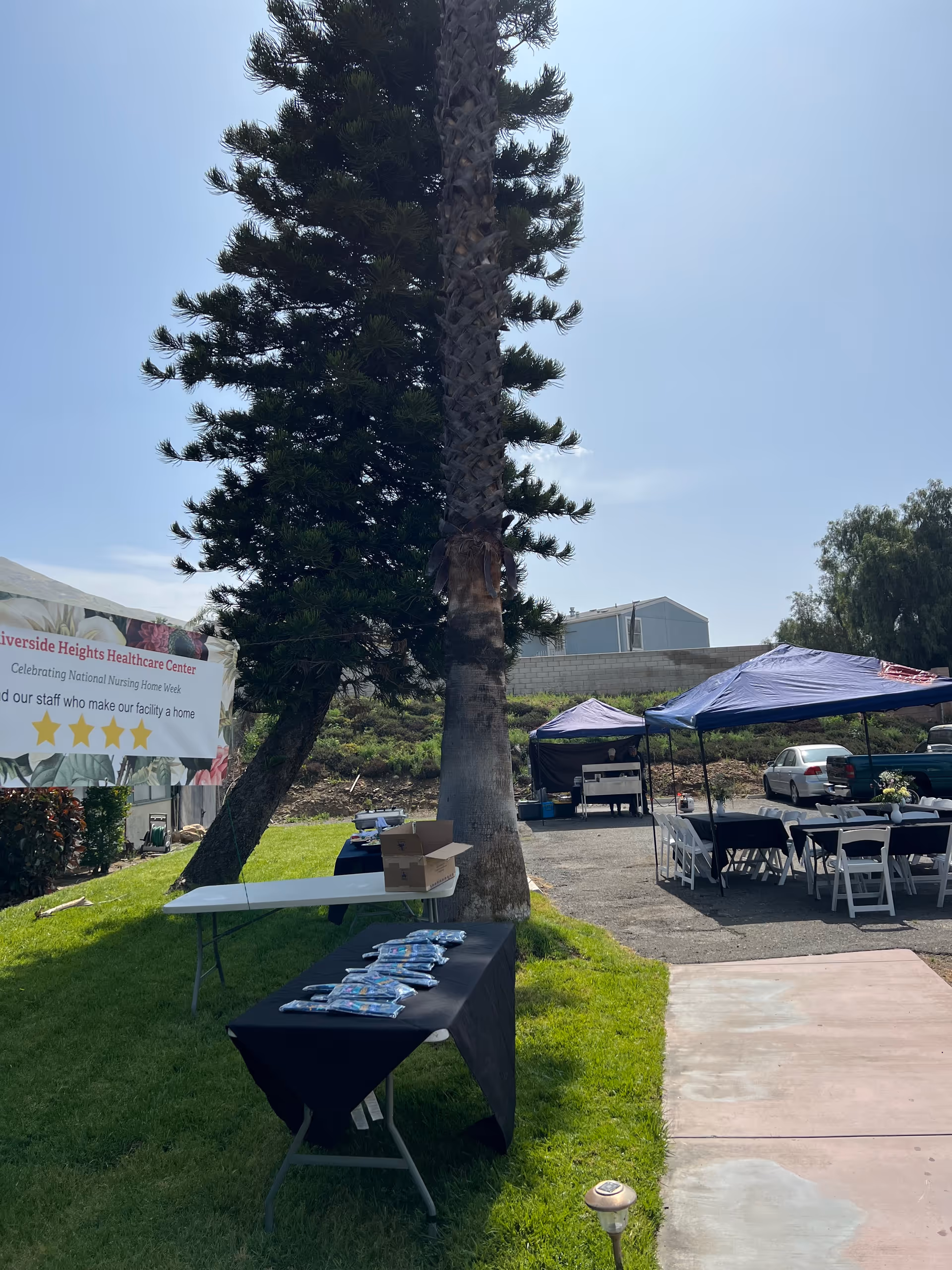 Outdoor event setup at Riverside Heights Healthcare Center with tables covered in black and white tablecloths, tents providing shade, a palm tree, and a banner celebrating National Nursing Home Week. There are chairs arranged around tables and some items placed on the tables.