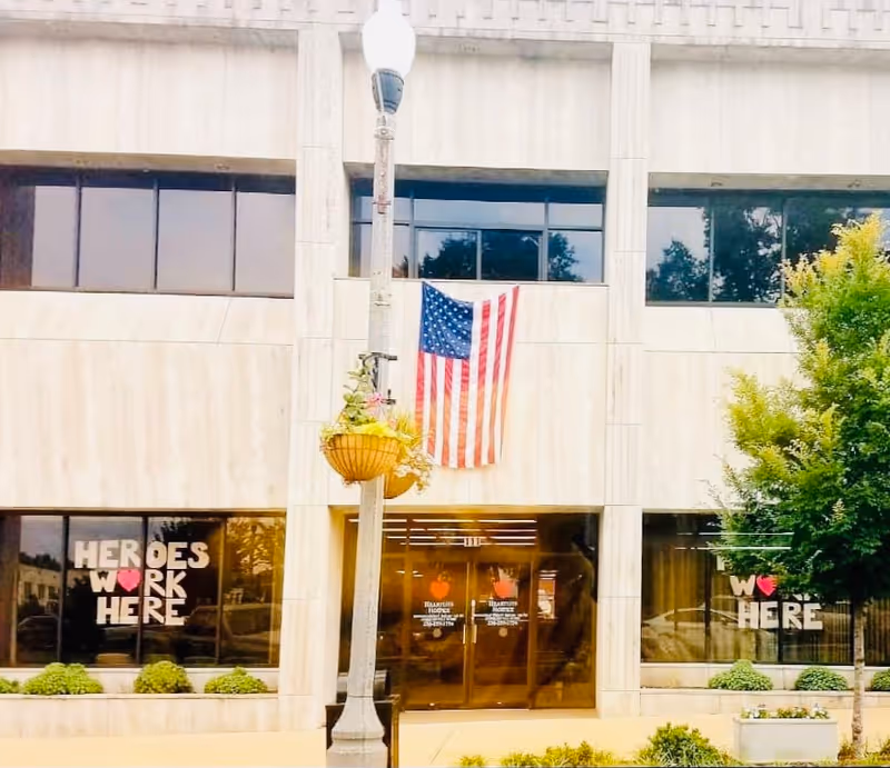 Front of Heartlite Hospice Alabama building with flag, glass doors, and hanging planter.