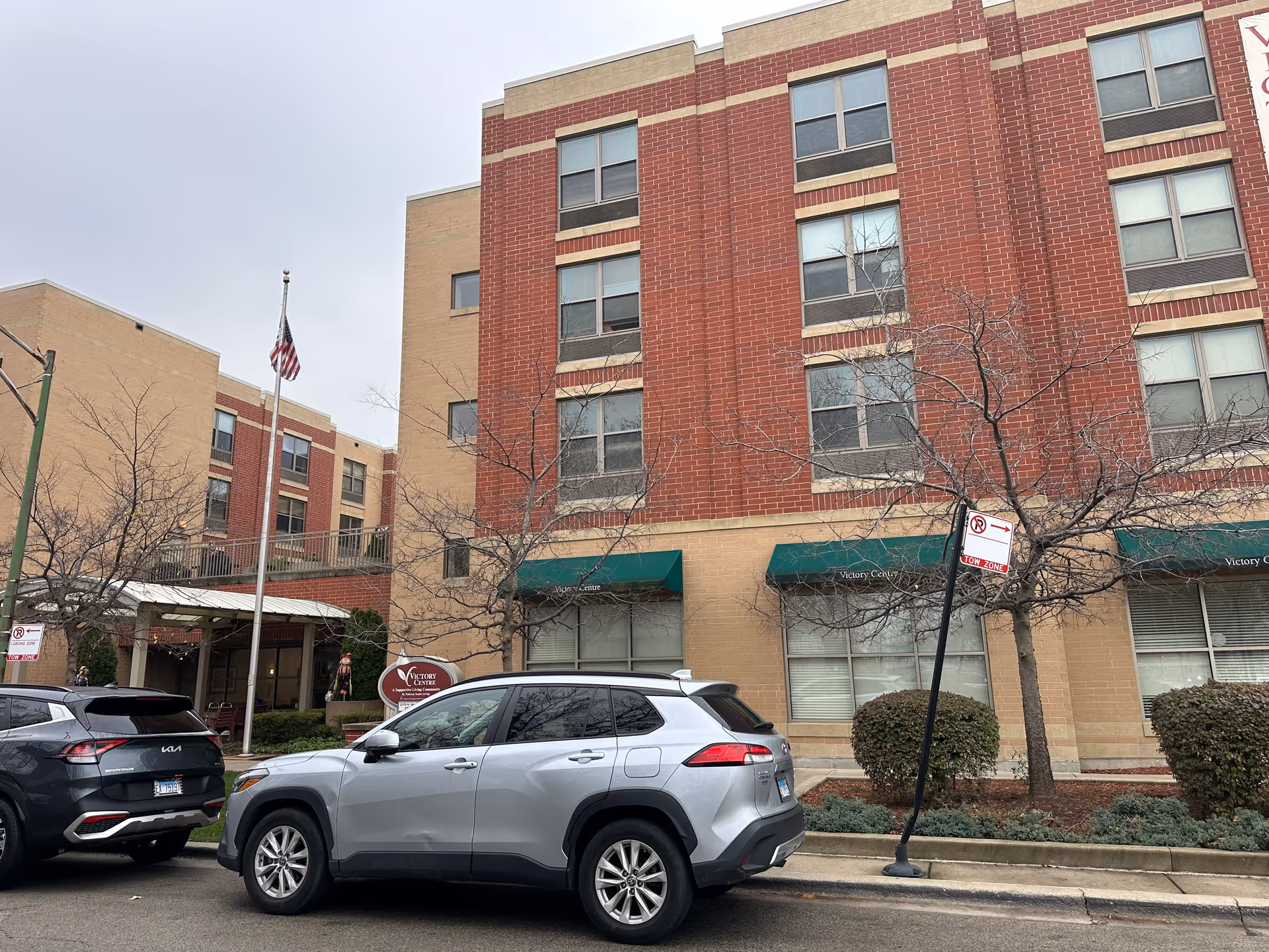 Exterior view of a multi-story senior living facility named Victory Centre of Galewood with brick and beige walls, several windows, leafless trees, and two parked cars in front. An American flag is visible on a flagpole near the entrance.