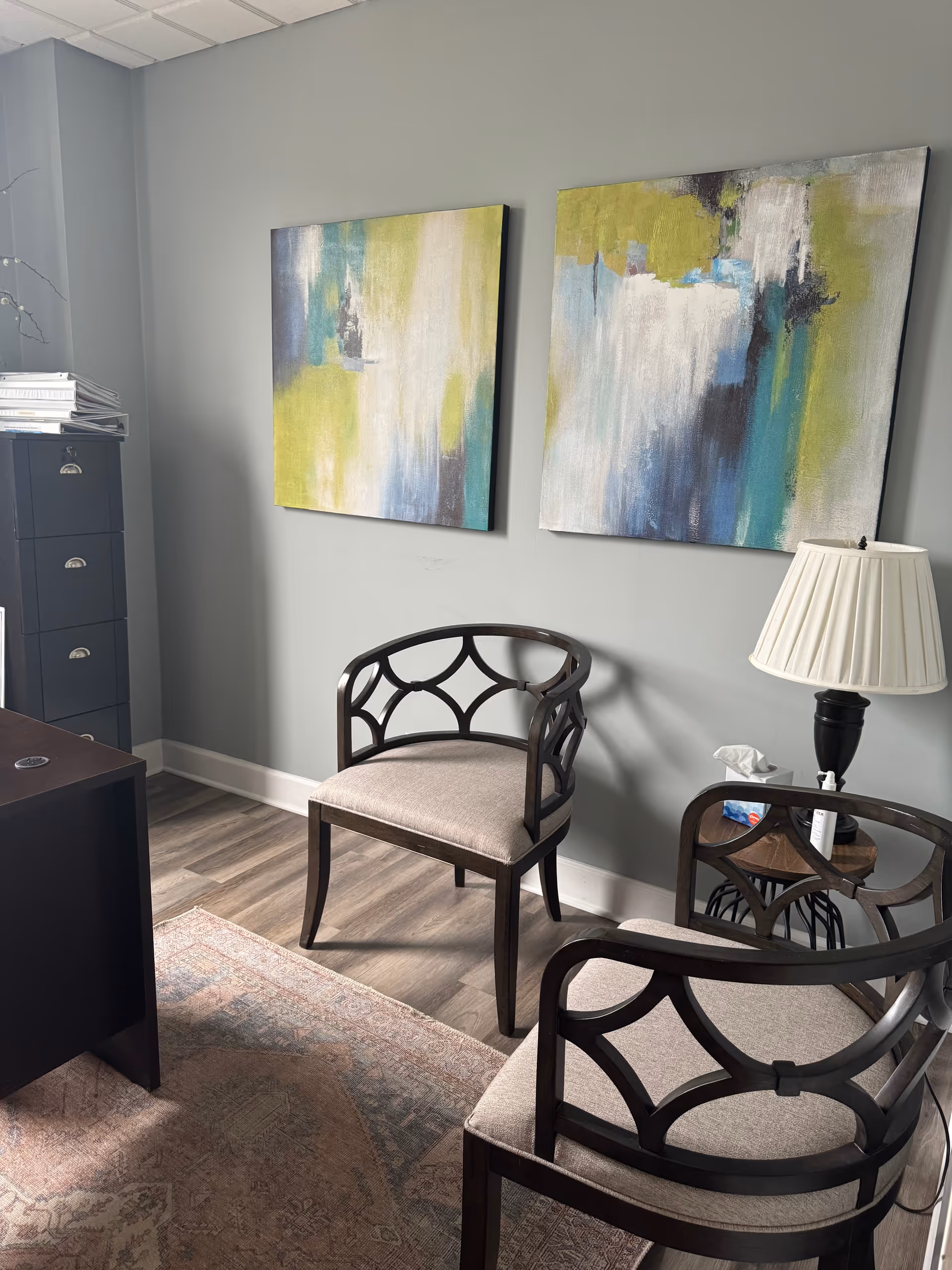 Interior corner with abstract wall art, two chairs, side table, lamp, cabinet, and rug.