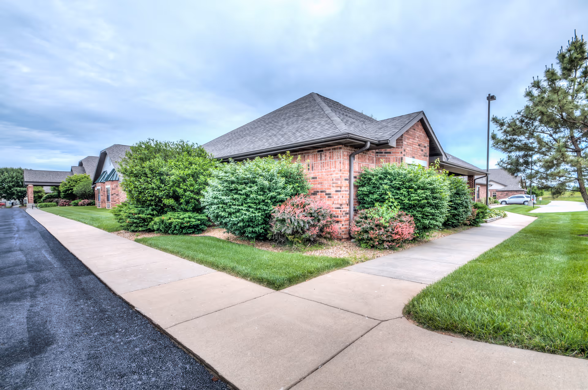Exterior view of a brick building with a pitched roof surrounded by well-maintained green bushes and grass. Sidewalks run along both sides of the building, with a paved road on the left and a grassy area with a tree on the right under a cloudy sky.