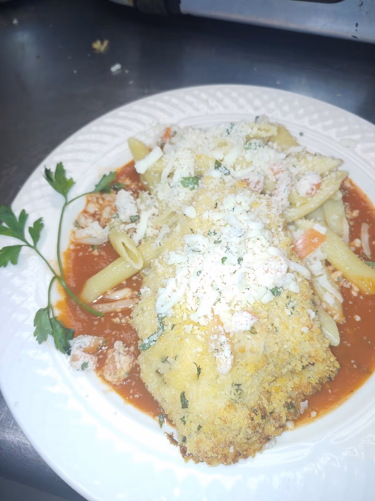 A white plate with a serving of baked breaded fish topped with shredded cheese and herbs, placed on a bed of penne pasta mixed with diced vegetables and marinara sauce, garnished with a sprig of parsley.