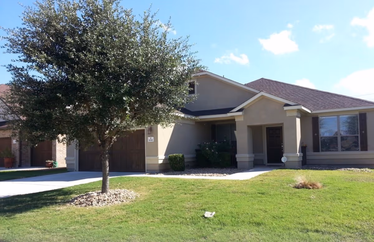Front exterior of a single-story beige assisted living home with a tree and lawn.