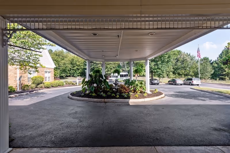 Covered porte-cochère entrance with a central planted island, parked cars, and an American flag in front of the building.