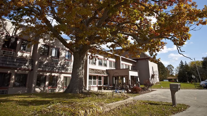 Front exterior of a two-story assisted living building with a large tree on the lawn and a mailbox by the driveway in autumn.