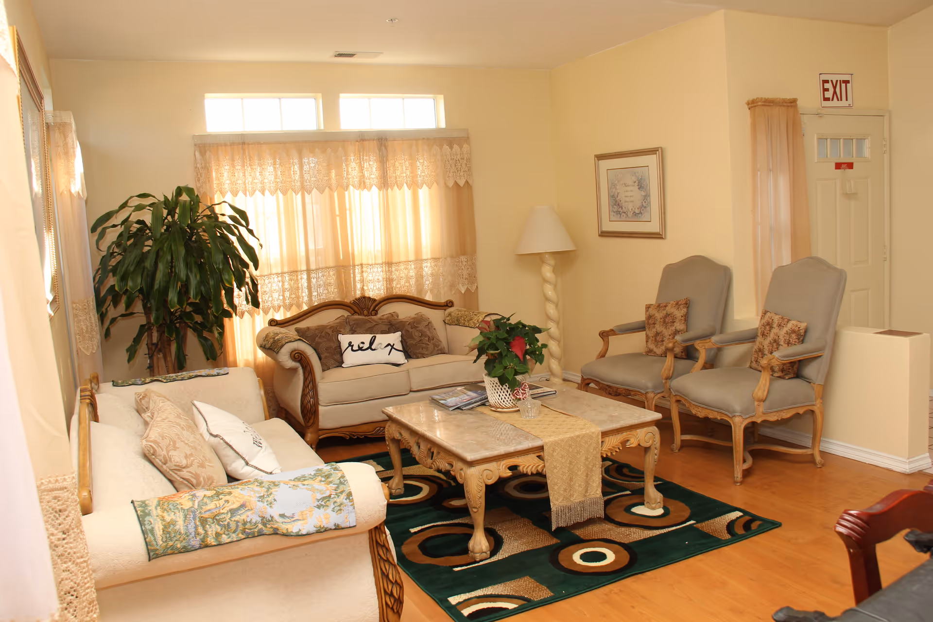 A cozy living room with two beige sofas adorned with decorative pillows, two upholstered armchairs with floral cushions, a wooden coffee table with a beige runner and a potted plant, a tall floor lamp, framed artwork on the wall, lace curtains on the windows, and a green patterned rug on a wooden floor. An exit door is visible in the background.