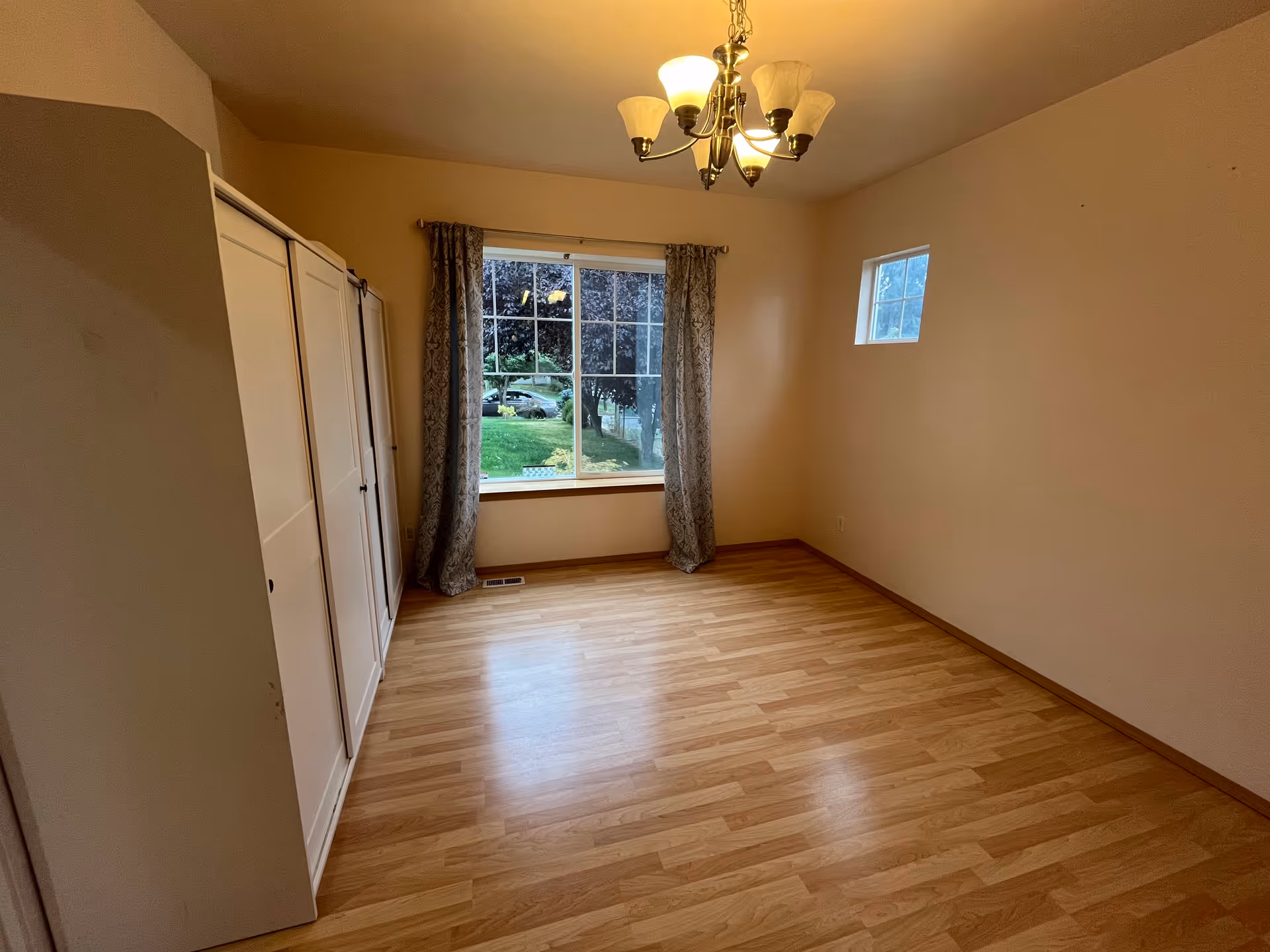 An empty bedroom with white wardrobes, a window, curtains, and a chandelier.