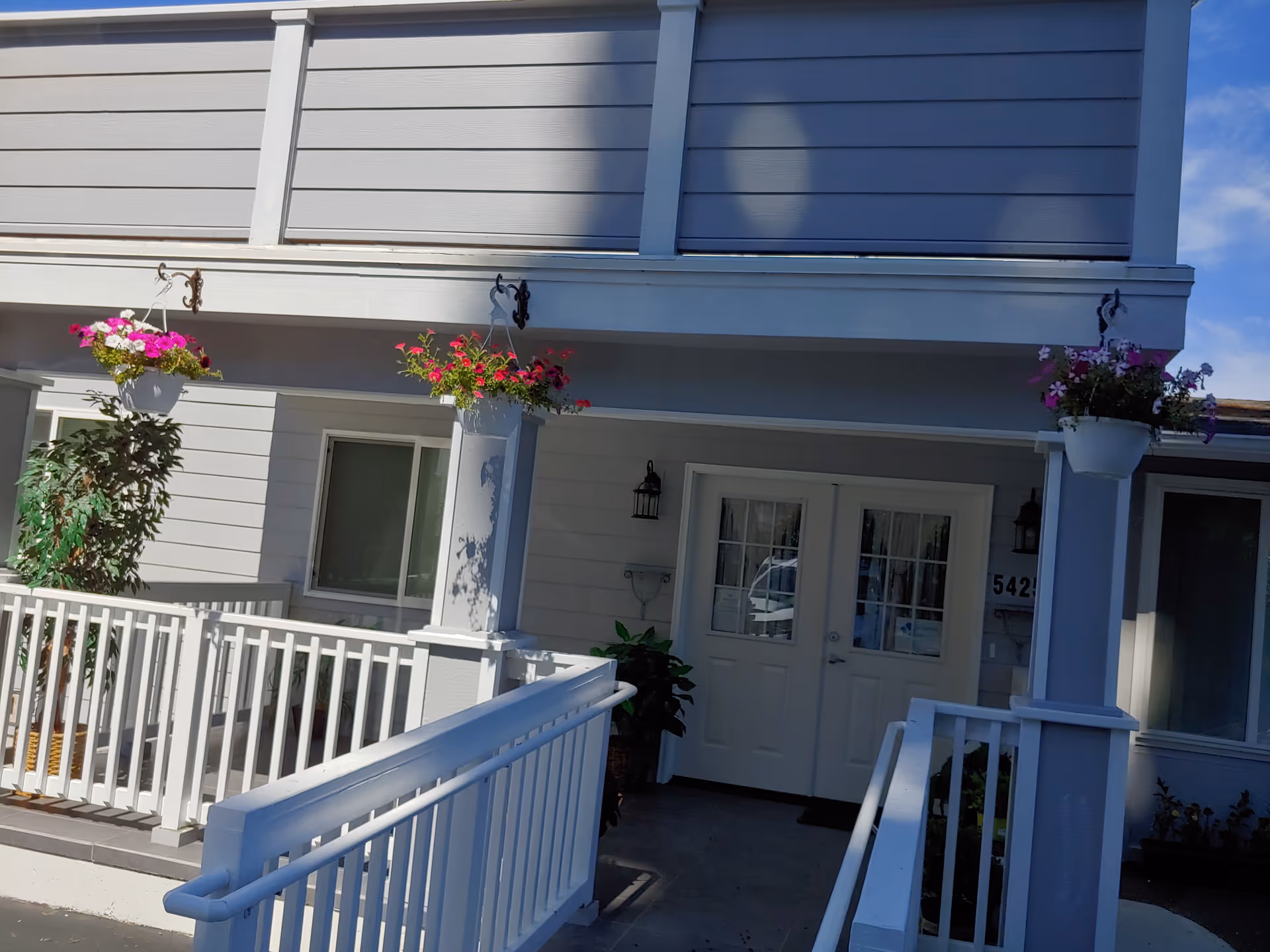 Front entrance of a light-gray senior living building with white doors and hanging flower baskets
