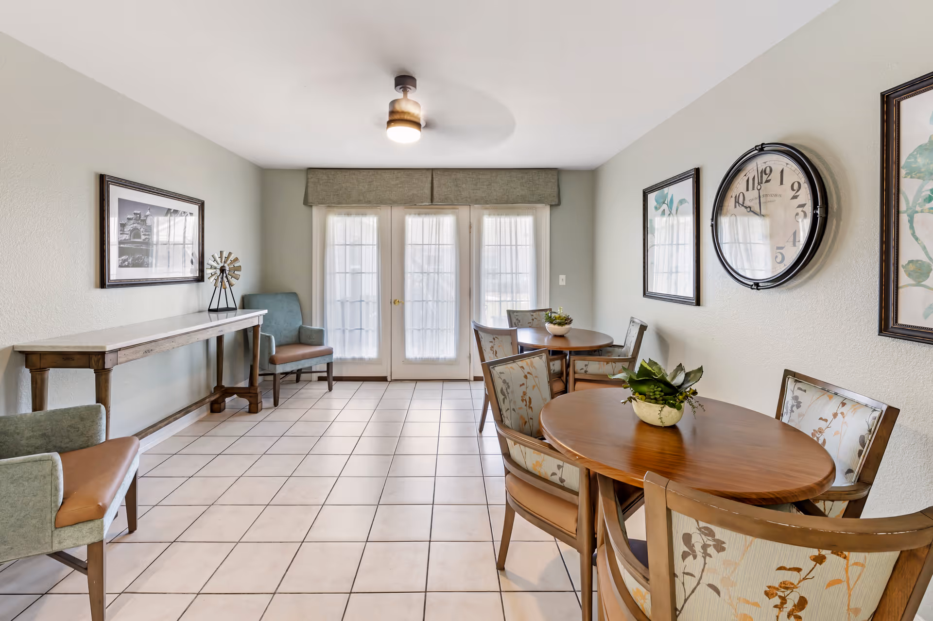 Bright communal dining area with round wooden tables and upholstered chairs, tile floor, wall clock, and French doors.