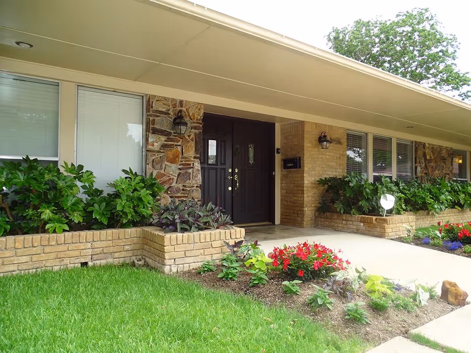 Front entrance of a single-story brick building with a covered porch, dark double doors, and landscaped flower beds.