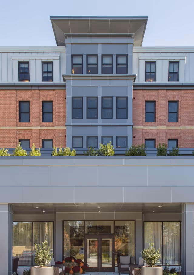 Modern multi-story senior living facility entrance with glass doors and planters.