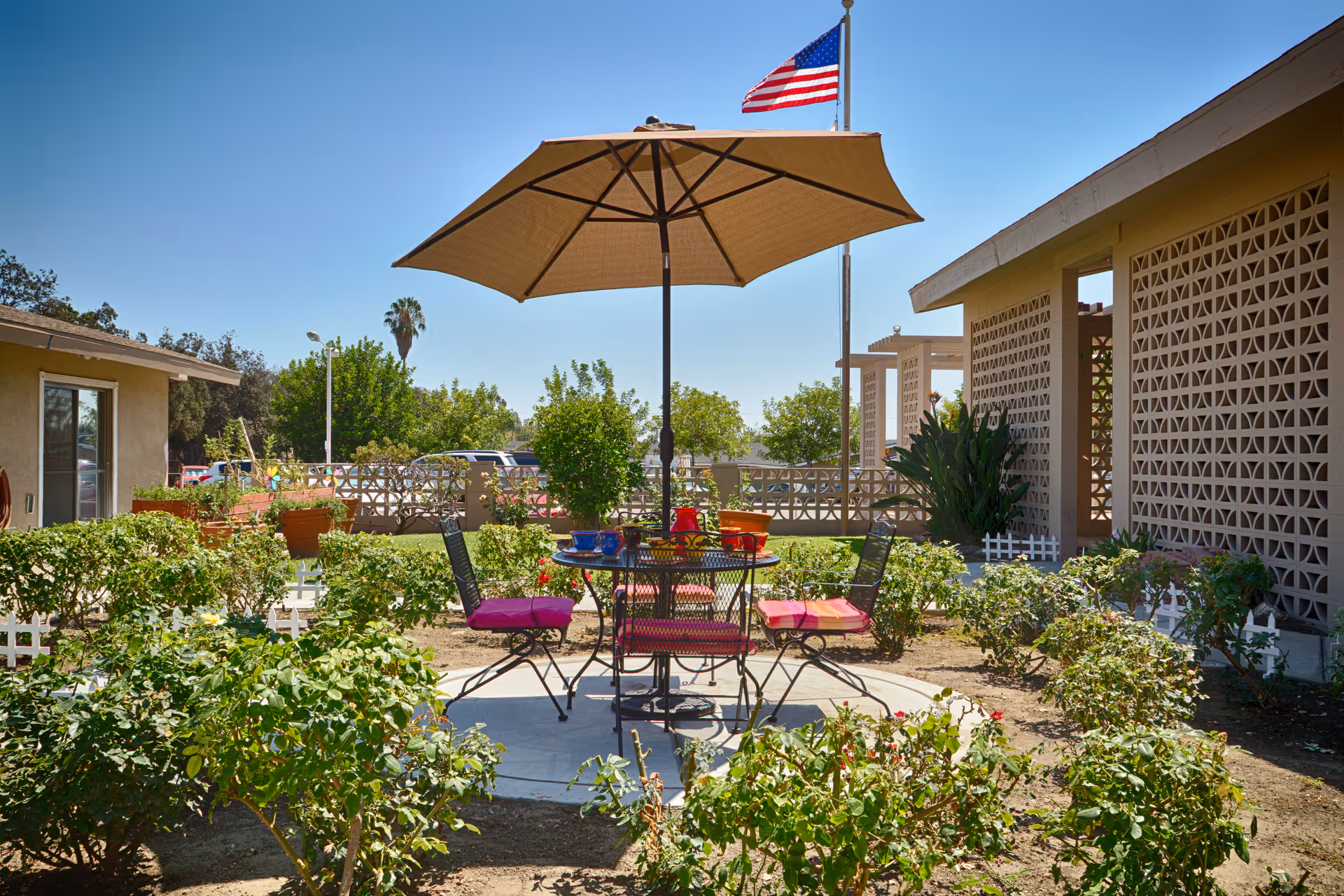 Sunny courtyard patio with a table, umbrella and chairs surrounded by shrubs and nearby buildings and an American flag.