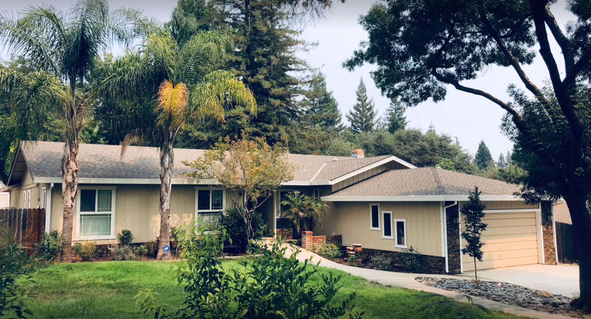 Single-story ranch-style house with a front lawn, driveway leading to a two-car garage and palm trees.