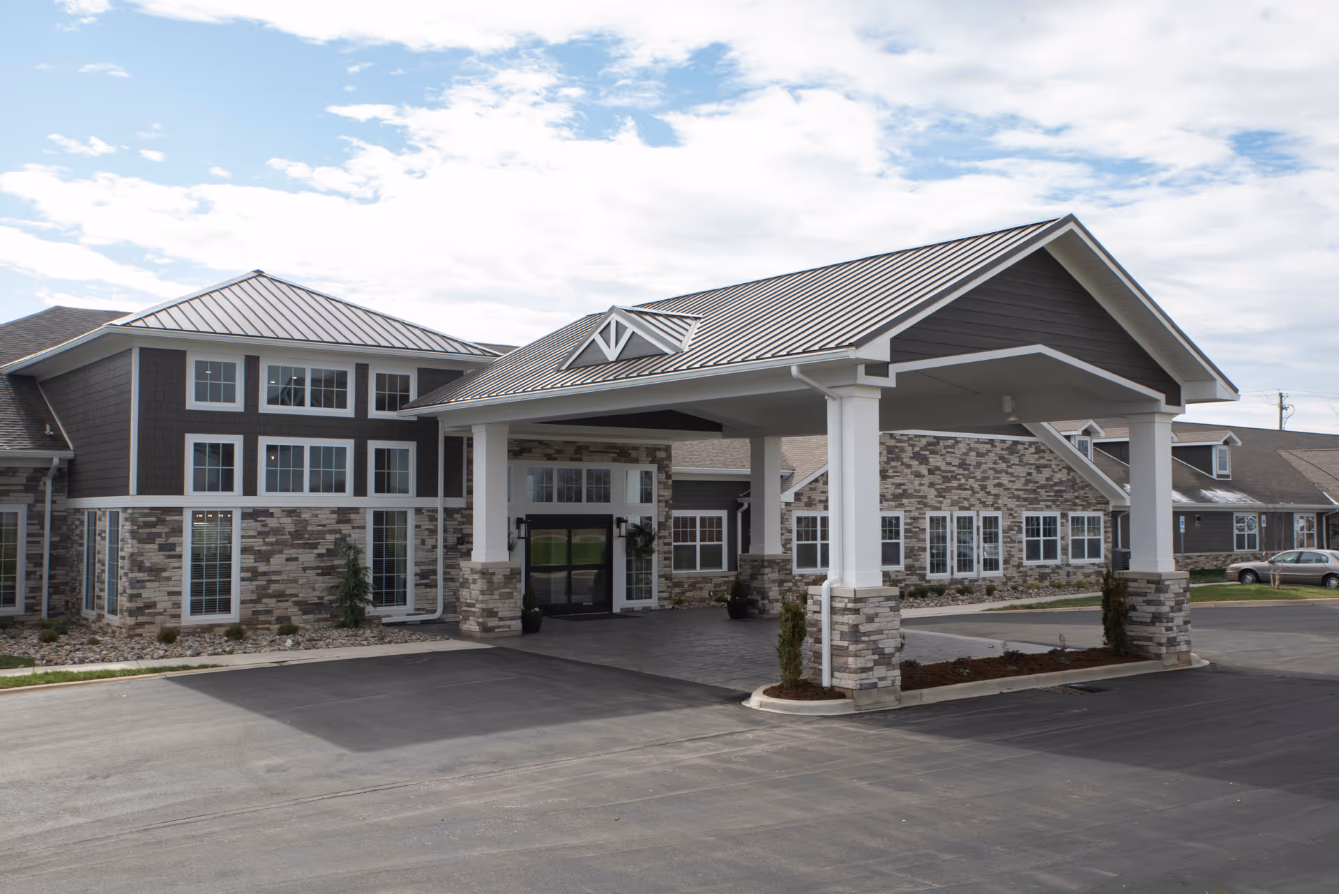 Exterior view of Boonesboro Trail Senior Living facility showing the main entrance with a covered drop-off area supported by white columns and stone accents. The building features a combination of stone and dark siding with multiple windows and a metal roof under a partly cloudy sky.