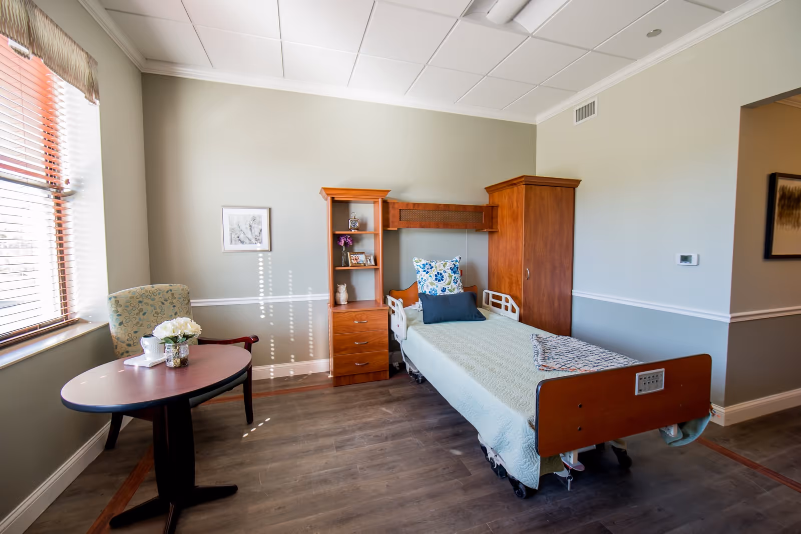 A bright, furnished senior bedroom with a single adjustable bed, wooden storage units, and a round table and chair by a window.