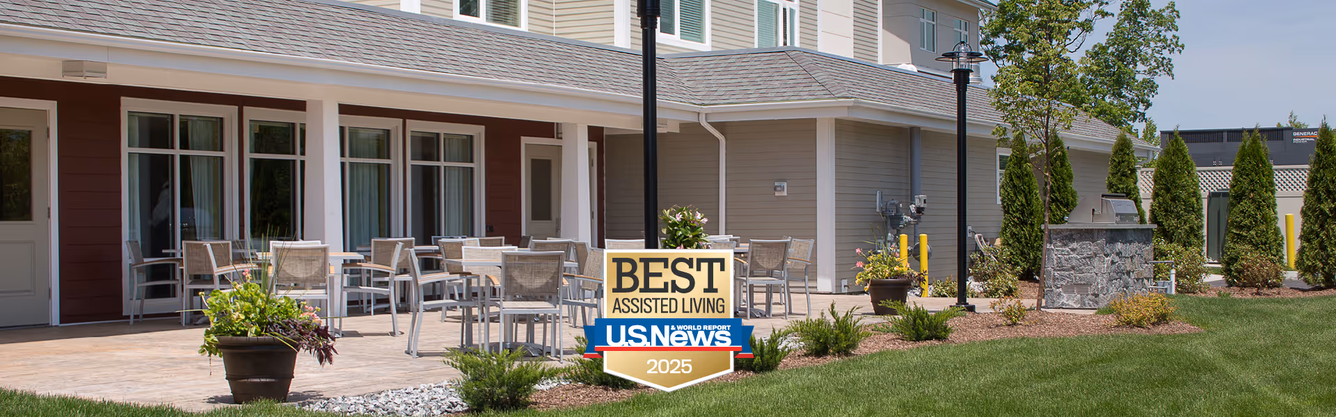 Outdoor patio area at Benchmark Senior Living at Split Rock featuring several tables and chairs on a paved surface, potted plants, and a landscaped garden with shrubs and trees. A sign in the foreground reads 'Best Assisted Living U.S. News 2025'. The building exterior is visible with multiple windows and doors.