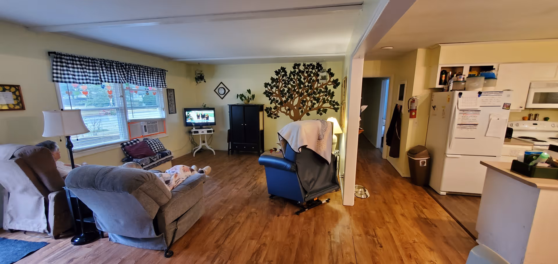 A cozy living room area in an assisted living facility with two elderly people seated in recliners facing a small TV on a stand. The room has wooden flooring, a window with checkered valance curtains, a wall decorated with a large tree mural, and a small kitchen area visible to the right with a refrigerator, stove, and microwave.