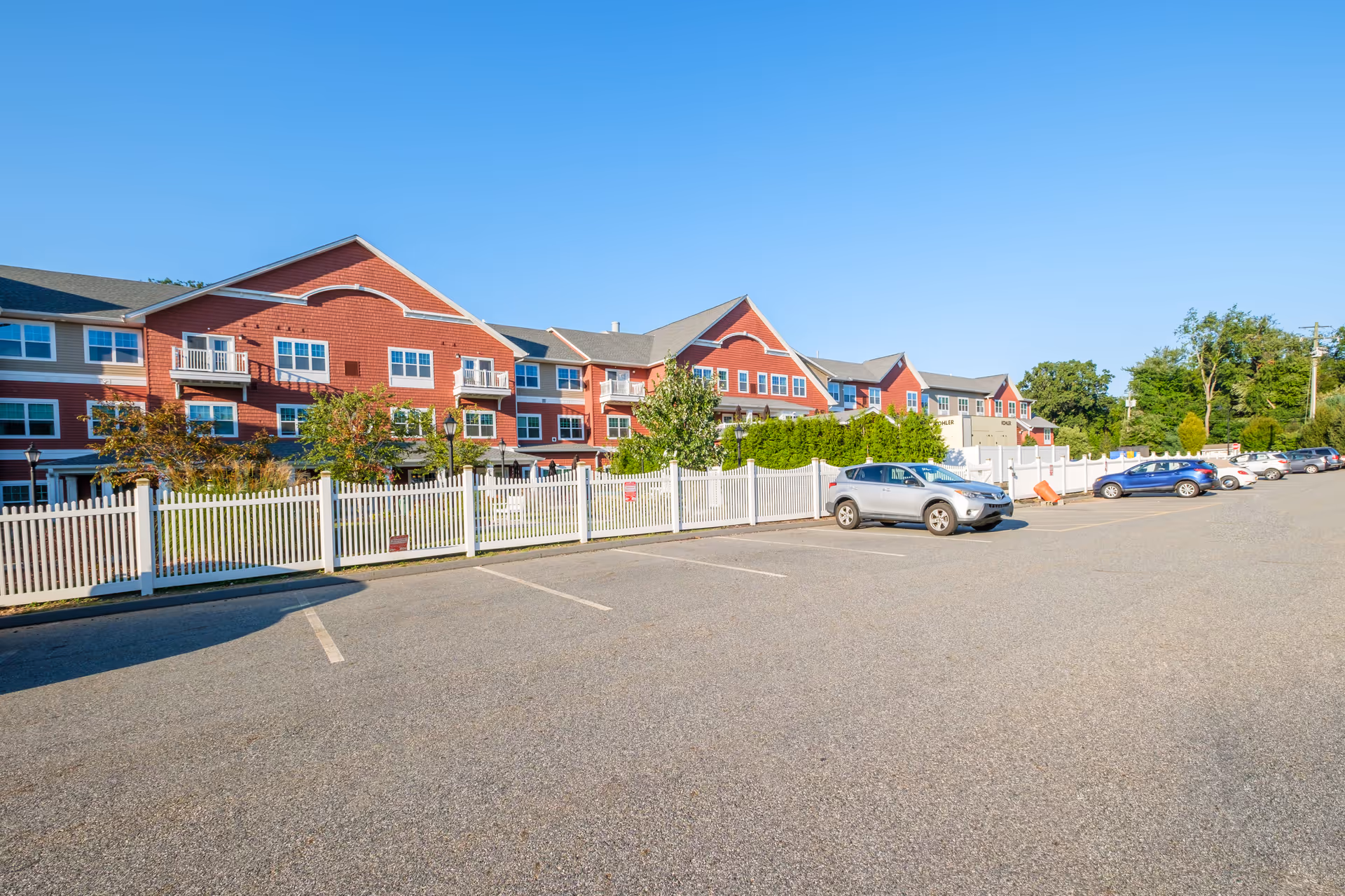 Parking lot and white picket fence in front of a large red multi-story senior living building with several parked cars under a clear blue sky.