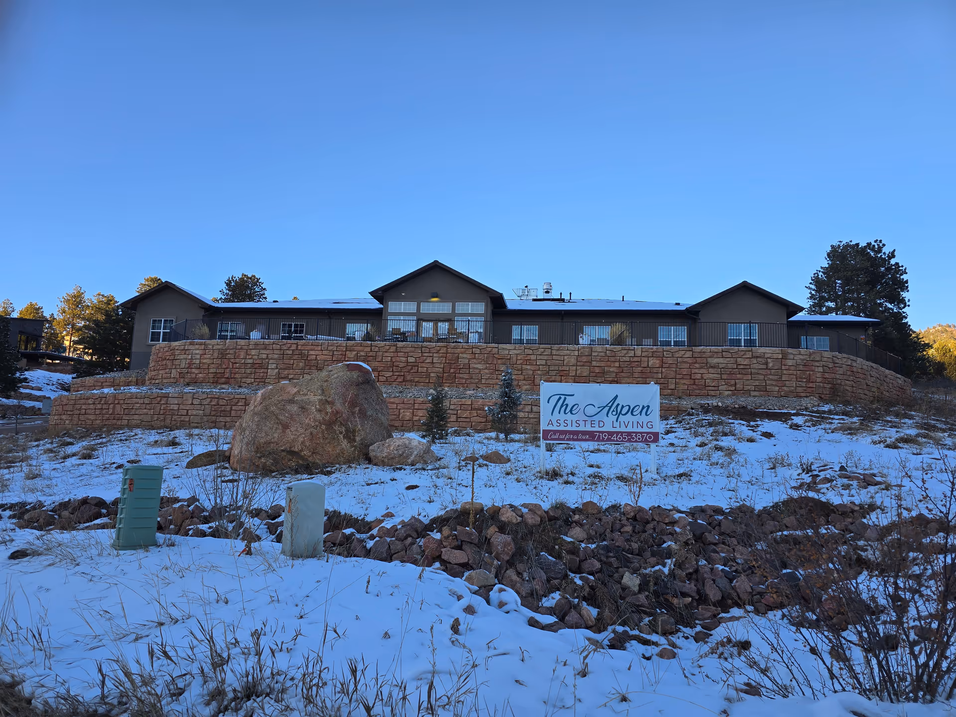 Exterior view of The Aspen assisted living facility on a snowy day, showing a single-story building with a stone retaining wall and a sign in front displaying the facility's name and contact number.