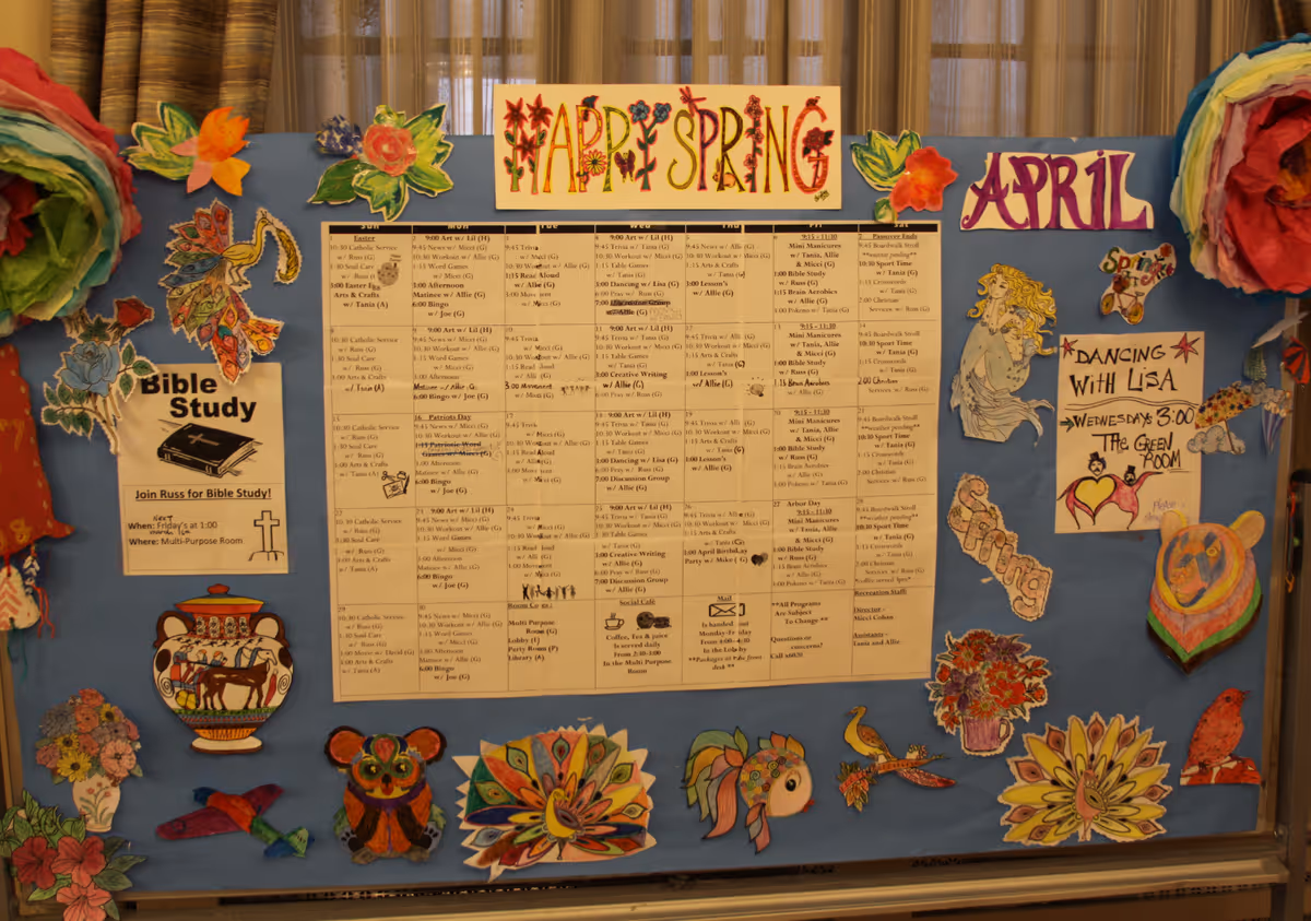 A decorated activity bulletin board showing an April schedule titled "Happy Spring" surrounded by colorful paper flowers and drawings.