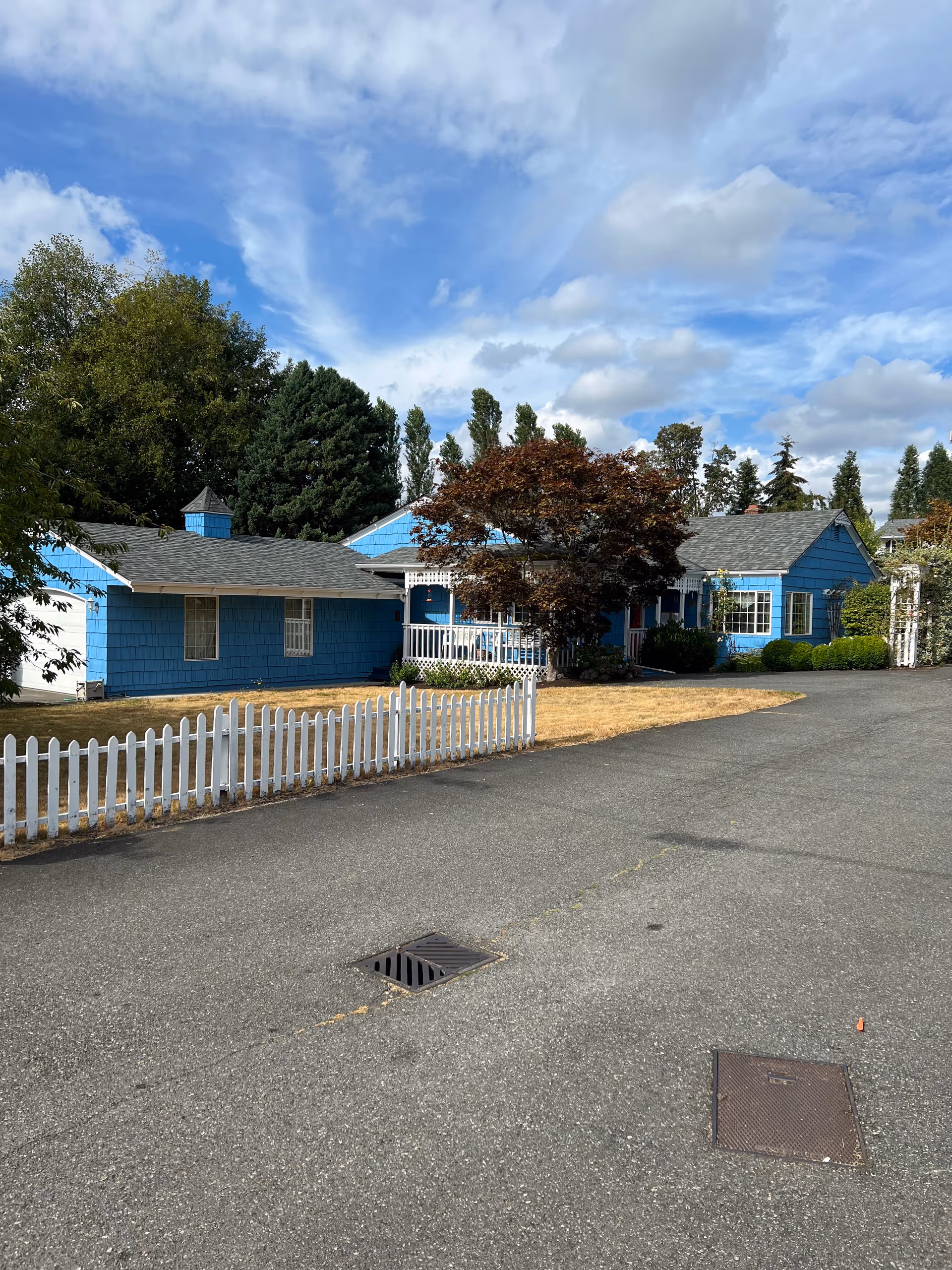 Blue single-story Birch Manor facility with white fence and driveway under cloudy sky