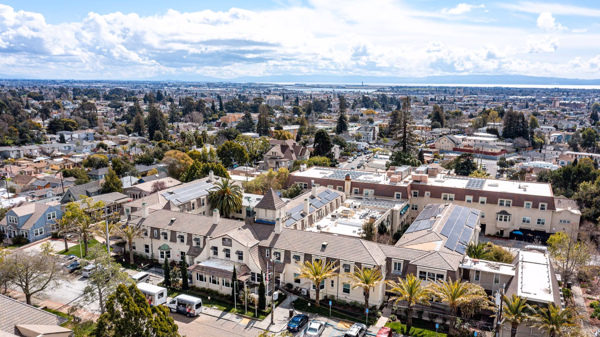 Aerial view of a large senior living complex with tiled roofs, palm trees, a parking area, and the surrounding residential neighborhood.