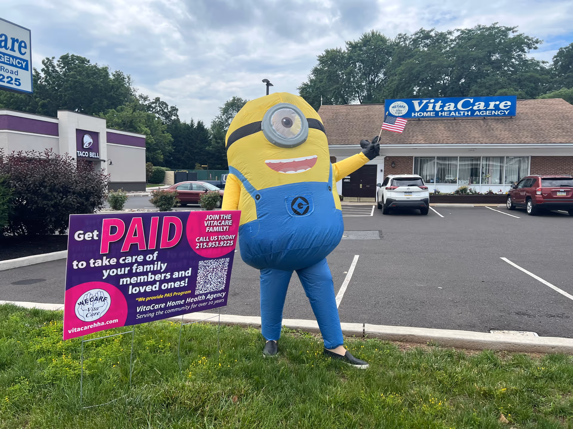 A large Minion costume stands outside VitaCare Home Health Agency with a parking lot and sign.